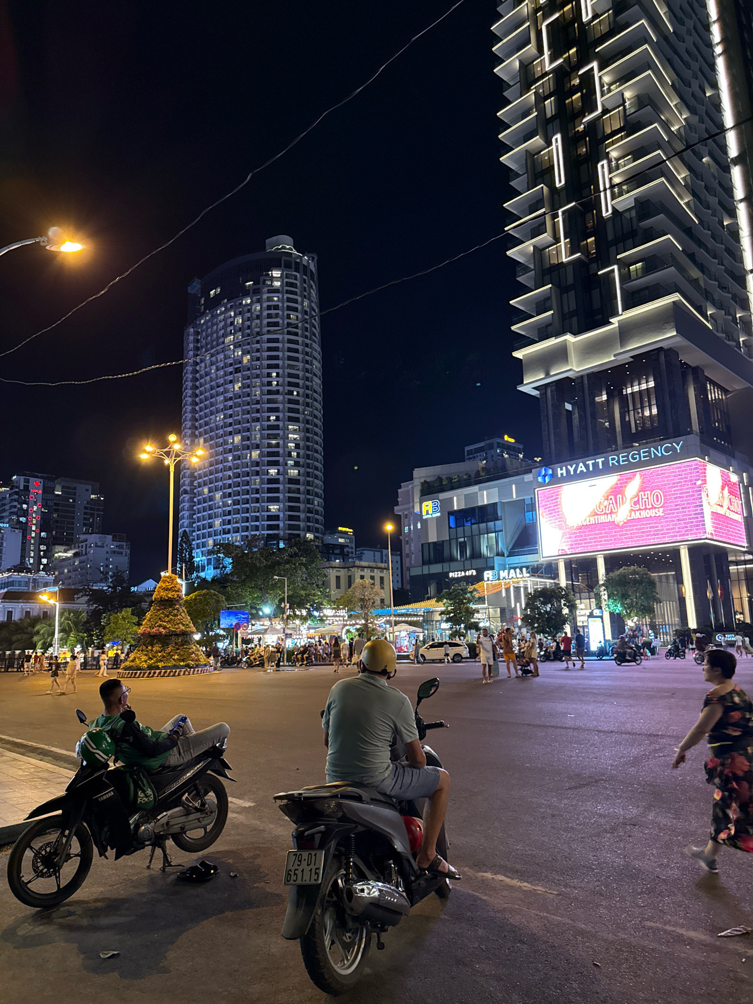 At night, in Nha Trang, Vietnam, a wide intersection is illuminated by streetlights and building lights. In the foreground, two men sit on motorcycles. The man on the left, wearing a green shirt and yellow helmet, is reclined on his motorcycle, looking to the left. His motorcycle is black, with "Yamaha" branding visible, and a green helmet rests on the seat. The man on the right, wearing a light green shirt and yellow helmet, faces away from the camera, looking ahead. His motorcycle is dark gray, and a portion of its license plate, "79-D1 851.15", is visible.
In the middle ground, a large, conical floral arrangement sits in the center of the intersection, surrounded by a low, circular wall. Pedestrians walk around the arrangement and in the background. Several streetlights with multiple bulbs are positioned around the intersection, casting a warm glow.
The background features several tall buildings, including a round, high-rise building with many lit windows and a modern skyscraper with a distinctive facade featuring horizontal and vertical white lighting elements. A large digital billboard displays "HYATT REGENCY" and "GALCHO ARGENTINIAN STEAKHOUSE" in bright pink and white. Other buildings with illuminated signs and windows are also visible. The sky is dark, indicating it is nighttime. The overall scene depicts a busy urban streetscape at night.