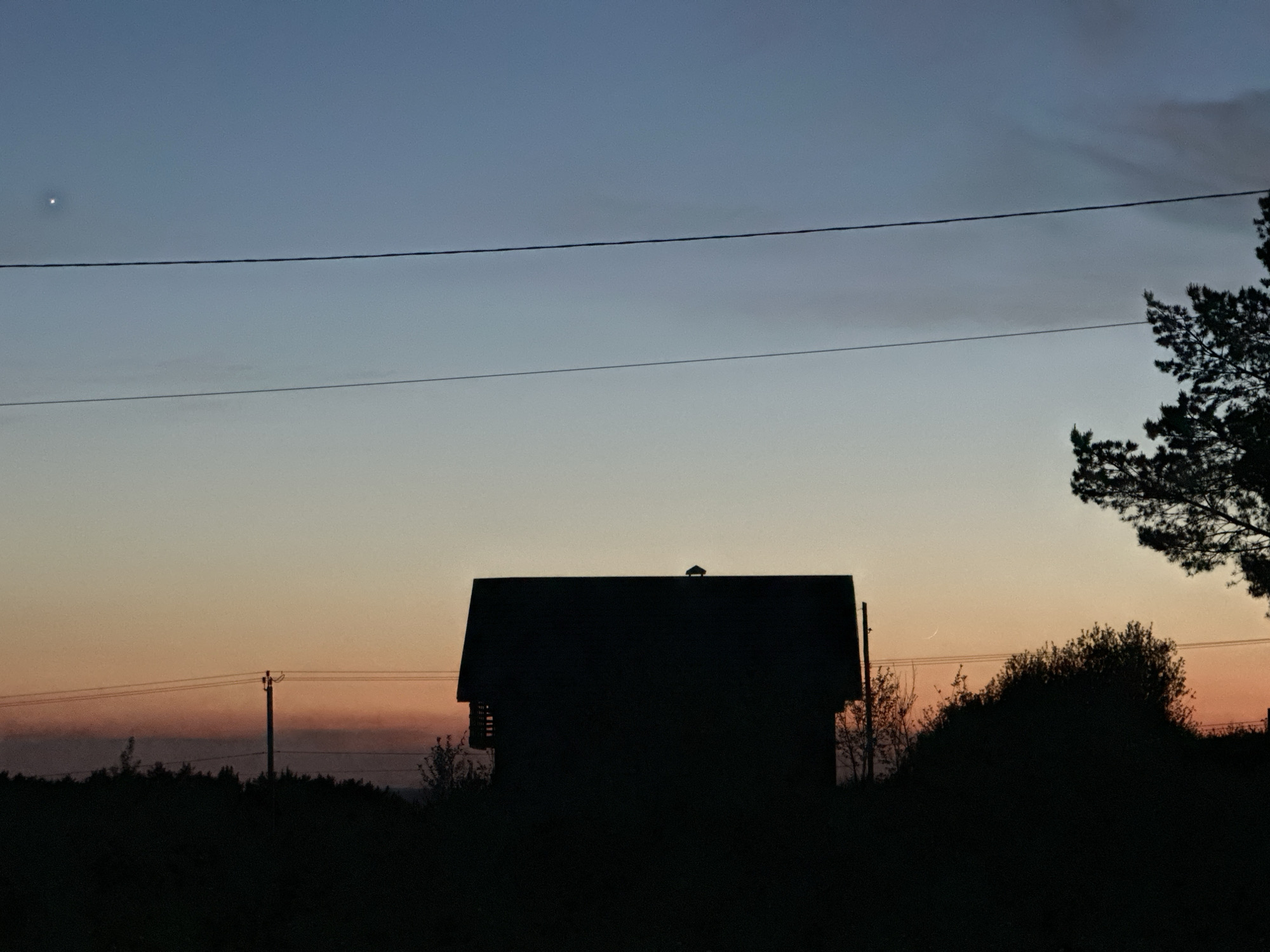 An outdoor shot depicts the silhouette of a house or building at dusk or twilight, likely located in the vicinity of Прикамский, Russia. The sky transitions from a light orange near the horizon to blue higher up. A bright spot, possibly a planet or star, is visible in the upper-left portion of the sky. Two power lines run horizontally across the frame. To the right of the house, there is a tree and some bushes, all silhouetted. A power pole is visible on the left side of the image. The foreground appears to be a field or another area with low vegetation.