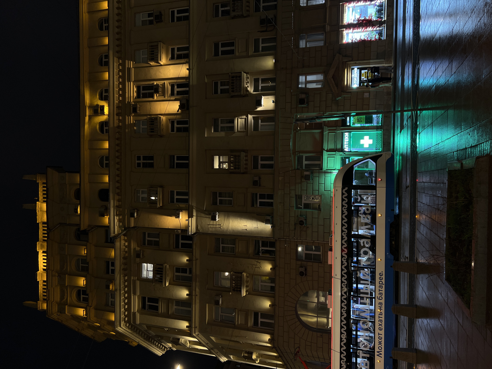 The image shows a multistory building at night with a light-colored facade. Many windows are lit, and air conditioning units are visible on the facade. A bright pharmacy sign with a green cross is visible on the ground floor. The street appears wet and reflective. A tram is partially visible in the foreground, with text in Russian on its side. The tram's color scheme is white and blue.