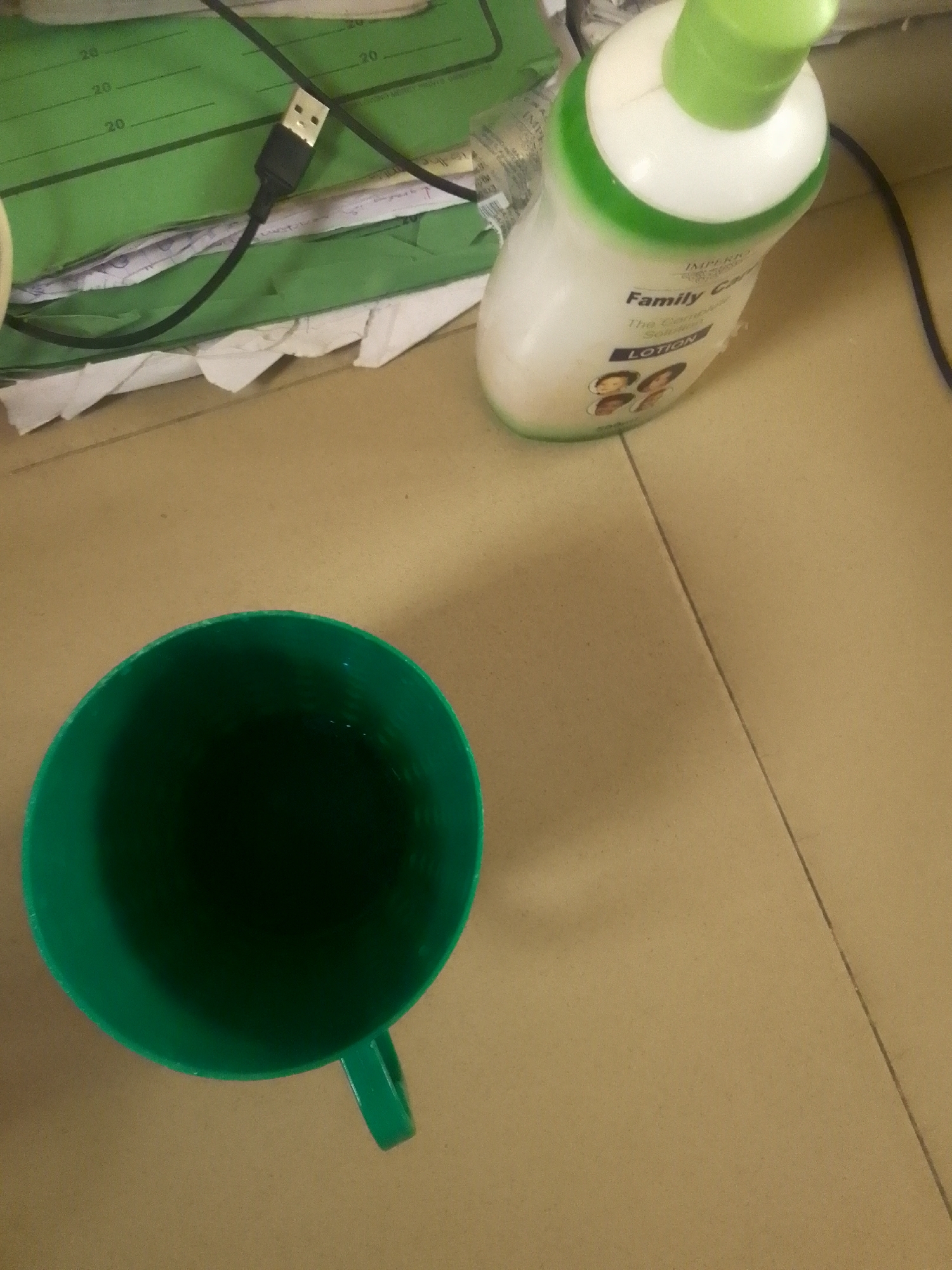The image shows a green plastic mug and a bottle of lotion placed on a light brown, tiled surface. A black USB cable is partially visible, connected to what appears to be a green folder with measurement markings. In the background, a stack of papers and the green folder are visible.
The lotion bottle has a white body with a green cap and a green band around its neck. Text on the bottle includes "IMPERI" and "Family Care LOTION." There are also images of four cartoon faces on the bottle.
The scene appears to be indoors, possibly on a desk or table. There are no explicit indicators of the time of day or weather. The overall impression is that of a casual, everyday setting, with items commonly found in a home or office. The arrangement of objects suggests a still life rather than an active event. No people are visible in the image.
The visible text on the lotion bottle is "IMPERI" and "Family Care LOTION." The green folder also has numerical markings that appear to be measurements, possibly in centimeters or inches.
Given the provided location context of Jalingo, Nigeria, this scene could be taking place in a home, office, or study area within that region.