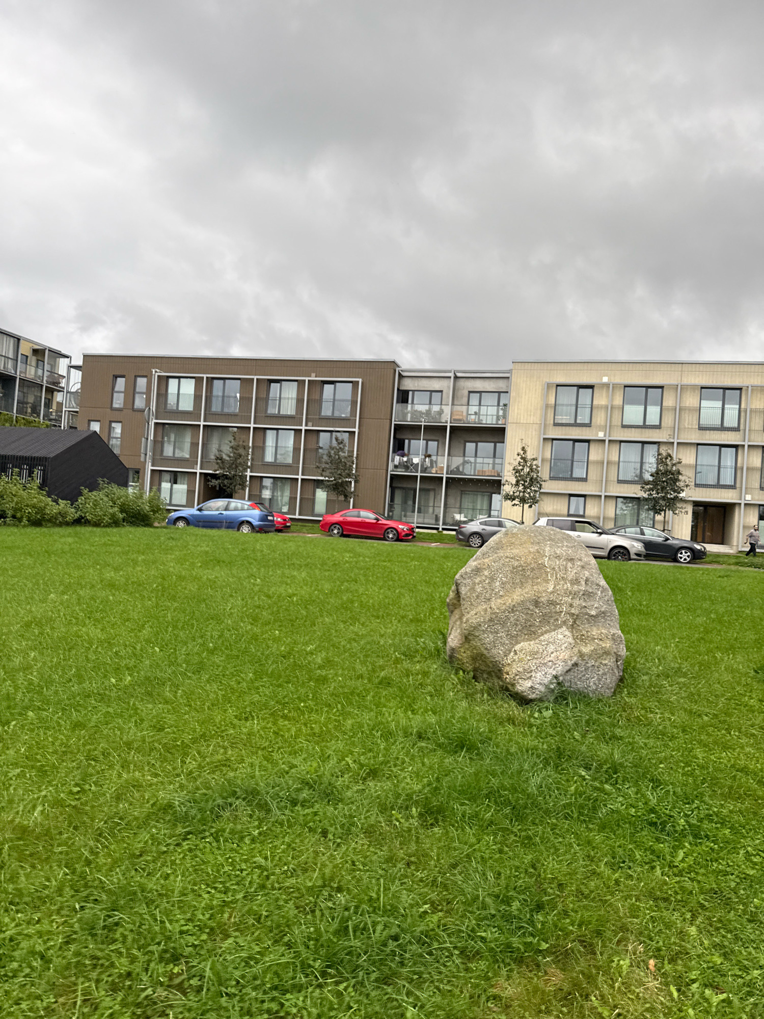 A medium-wide shot shows a park area in front of a row of modern apartment buildings under a cloudy sky. A large boulder is in the foreground, resting on a neatly mown green lawn. Behind the boulder, several cars are parked along the sidewalk in front of the apartment buildings. The buildings themselves are multi-story structures with light-colored facades and dark-framed windows. Some balconies are visible, and trees are planted between the buildings and along the sidewalk. To the left, a dark, angular building with a pitched roof is situated in front of one of the apartment buildings. The scene appears to be located in the Lasnamäe district of Tallinn, Estonia.