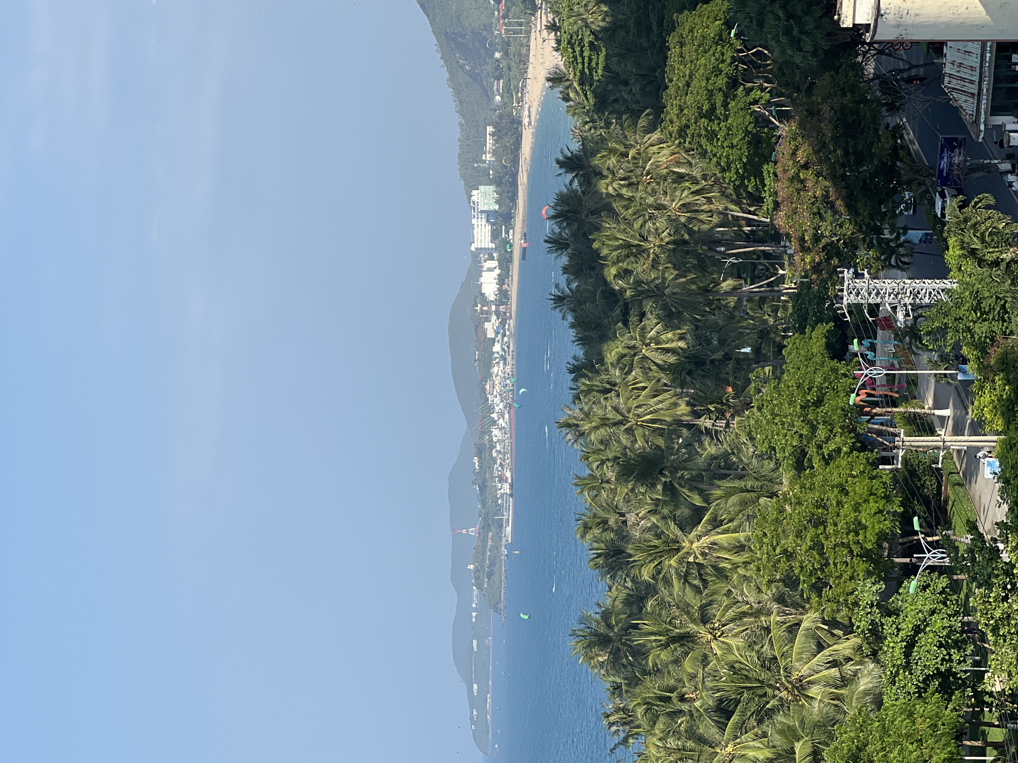 An aerial, wide-angle, high-angle shot captures a coastal scene in Nha Trang, Vietnam. The blue ocean dominates the upper half of the frame, dotted with several small boats and kite surfers. The coastline is lined with a sandy beach and a densely populated city featuring white and blue buildings. Lush green palm trees and other tropical vegetation fill the foreground and mid-ground, obscuring parts of a road with a few parked cars and a single pedestrian. The sky is clear and pale blue. The image appears to have been taken during daylight hours.