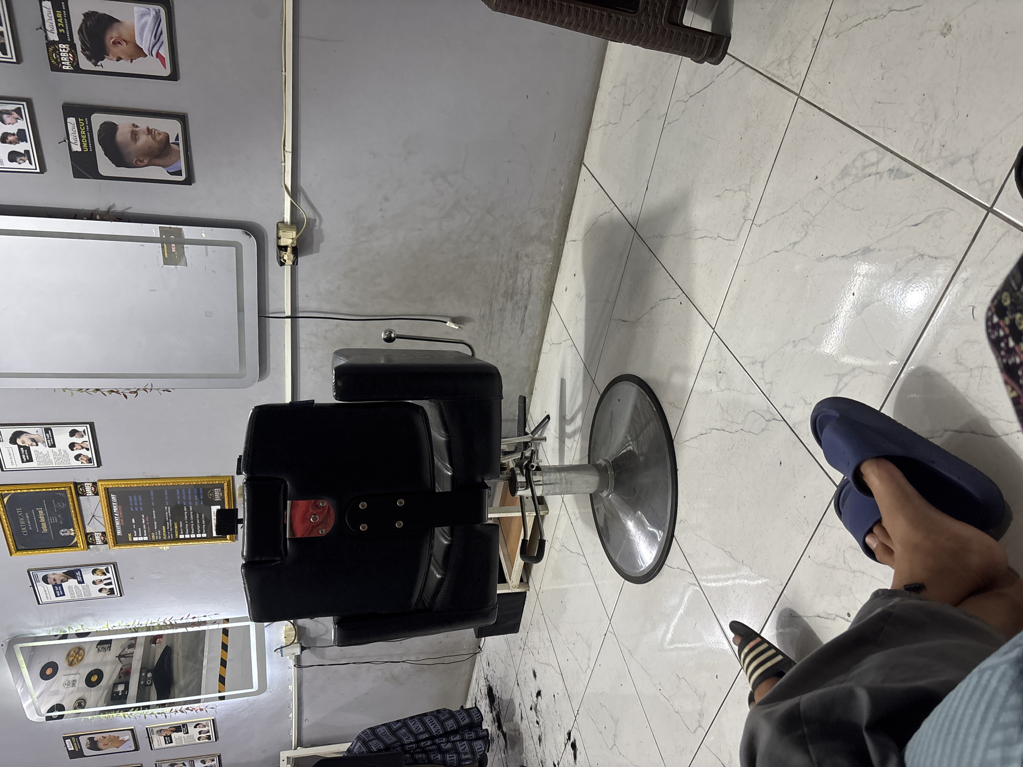 The image depicts the interior of a barbershop, likely located in Wanajaya, Indonesia. The scene is dominated by a black barber chair with a chrome base in the center. To the left of the chair is a large, illuminated mirror with a white frame. On the wall behind the chair, several framed pictures and certificates are displayed, showcasing various hairstyles and possibly barbershop achievements. One of these appears to be a price list for services offered. The floor is tiled in white with a marble pattern, and there are scattered hair clippings on the floor, indicating recent activity. In the foreground, two individuals are partially visible, wearing casual clothing and slippers, suggesting they are either customers or staff members relaxing. A brown plastic stool is visible on the right side of the frame. The overall environment suggests a functional and operational barbershop.