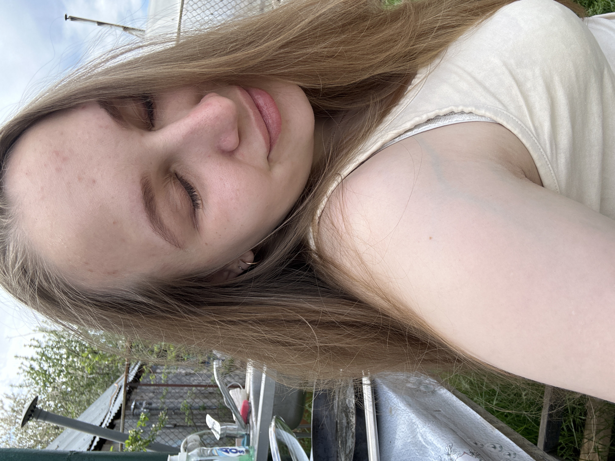 A young woman with long, light brown hair lies in an outdoor setting. Her eyes are closed, and she has fair skin with some blemishes on her forehead. She wears a light-colored tank top. The background includes greenery, a fence, and a building with a metal roof. The setting appears to be a garden or backyard. There is no information to suggest it is in Chaikovsky, Russia.