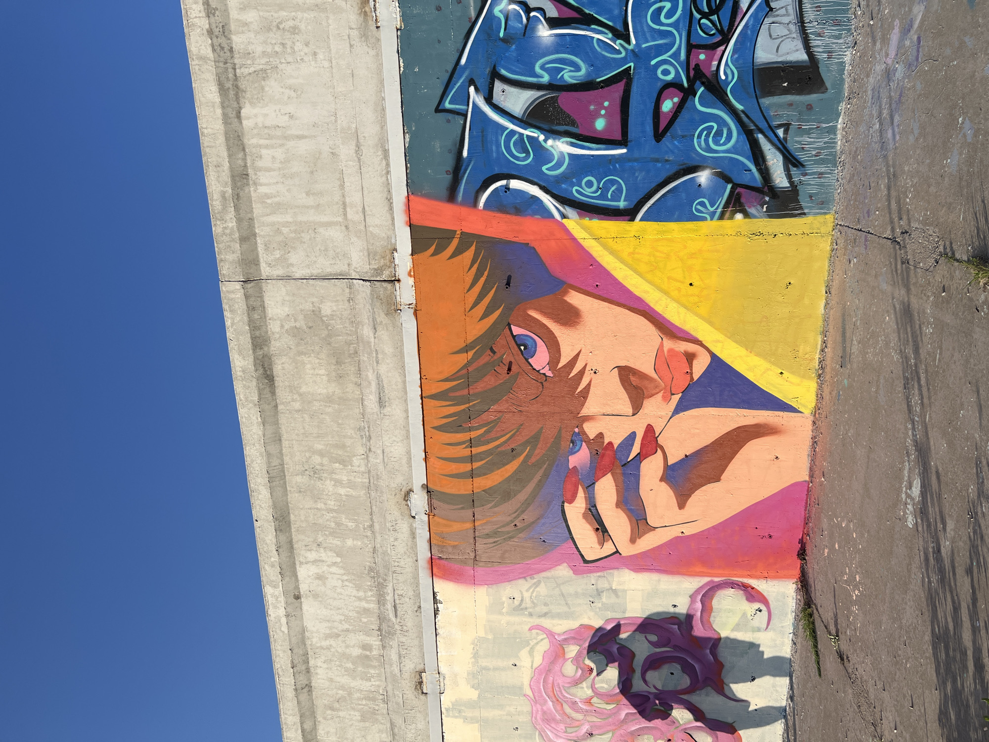 This is a photograph of graffiti on a concrete wall. The graffiti features a close-up of a person's face with orange hair and blue eyes, rendered in a stylized, anime-like manner. The person is wearing red nail polish and is partially covering their face with their hand. To the left of this central figure is a shadowy, purple and pink abstract design. To the right, there is blue graffiti with white and green lettering. The wall is situated outdoors, under a clear blue sky. The ground in front of the wall is concrete, with some sparse grass and debris. The concrete structure above the graffiti appears weathered and utilitarian. This scene is located in the city of Khabarovsk, Russia.