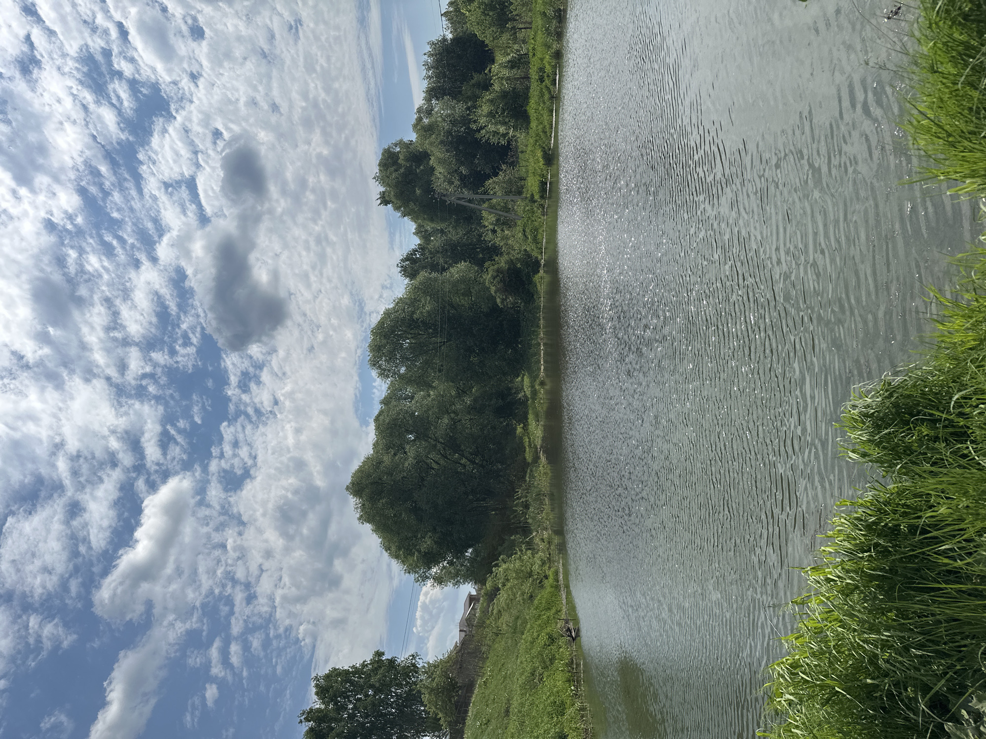 The image depicts a body of water, possibly a lake or wide river, with a shoreline lined by trees and grass. The water's surface is disturbed by gentle ripples. Above, the sky is filled with patchy clouds. The location is given as Эстачи, Russia, but there are no specific details visible that would identify this location. No people or objects are visible, only the natural environment.