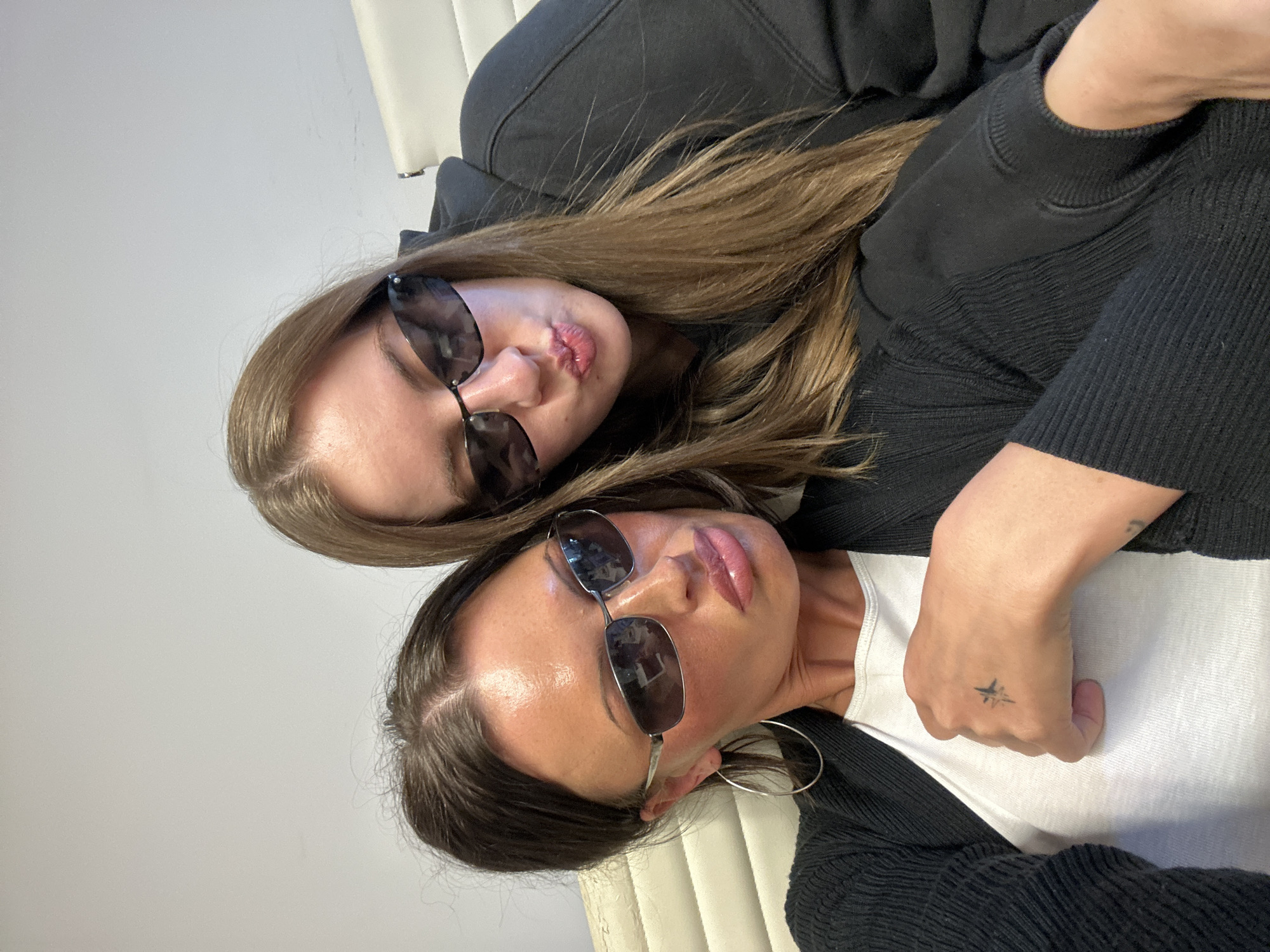 An indoor, close-up photograph features two young women, oriented ninety degrees clockwise. The women appear to be posing casually, with one embracing the other. The woman positioned towards the top of the frame has long, light brown hair and is wearing a dark hoodie. She is making a "duck face" or puckered lips expression and wears dark, rectangular sunglasses. The woman towards the bottom of the frame has darker brown hair, a neutral expression with slightly parted lips, and wears aviator-style sunglasses. She is dressed in a white t-shirt layered under a dark, possibly ribbed, cardigan or jacket. A large silver hoop earring is visible on her left ear, and a small, black star-shaped tattoo adorns the back of her left hand. The women are positioned against a plain, light-colored wall, with parts of what appears to be a light-colored chair or furniture visible in the background. The lighting suggests an indoor setting, and no specific time of day or weather can be determined from the image. There is no visible text in the image. The location context provided is London, United Kingdom.