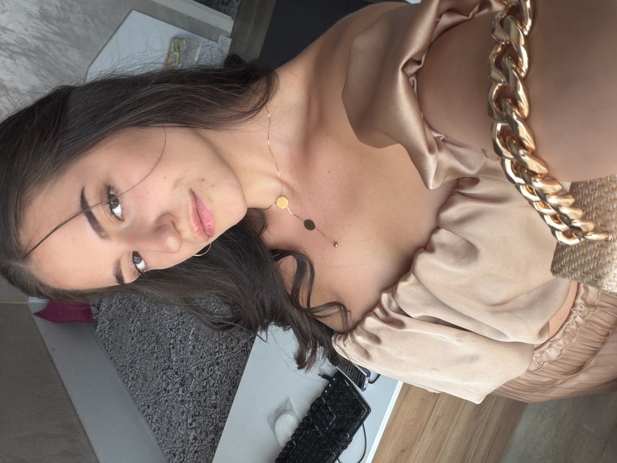 A young woman with long, dark hair and light brown eyes looks at the camera, her head tilted slightly. She is wearing a beige, off-the-shoulder top with a ruffled neckline and a gold chain bracelet on her right arm. A thin gold necklace with two circular pendants rests on her collarbone. Her makeup is subtle, with emphasis on her eyes and lips.
In the background, a desk is visible with a black keyboard and a small stack of white circular pads. A patterned rug covers a portion of the floor behind the desk, and a pink cushion is partially visible. The overall lighting suggests an indoor setting during the daytime. There is no specific indication of the location within the city of Burgas, Bulgaria.