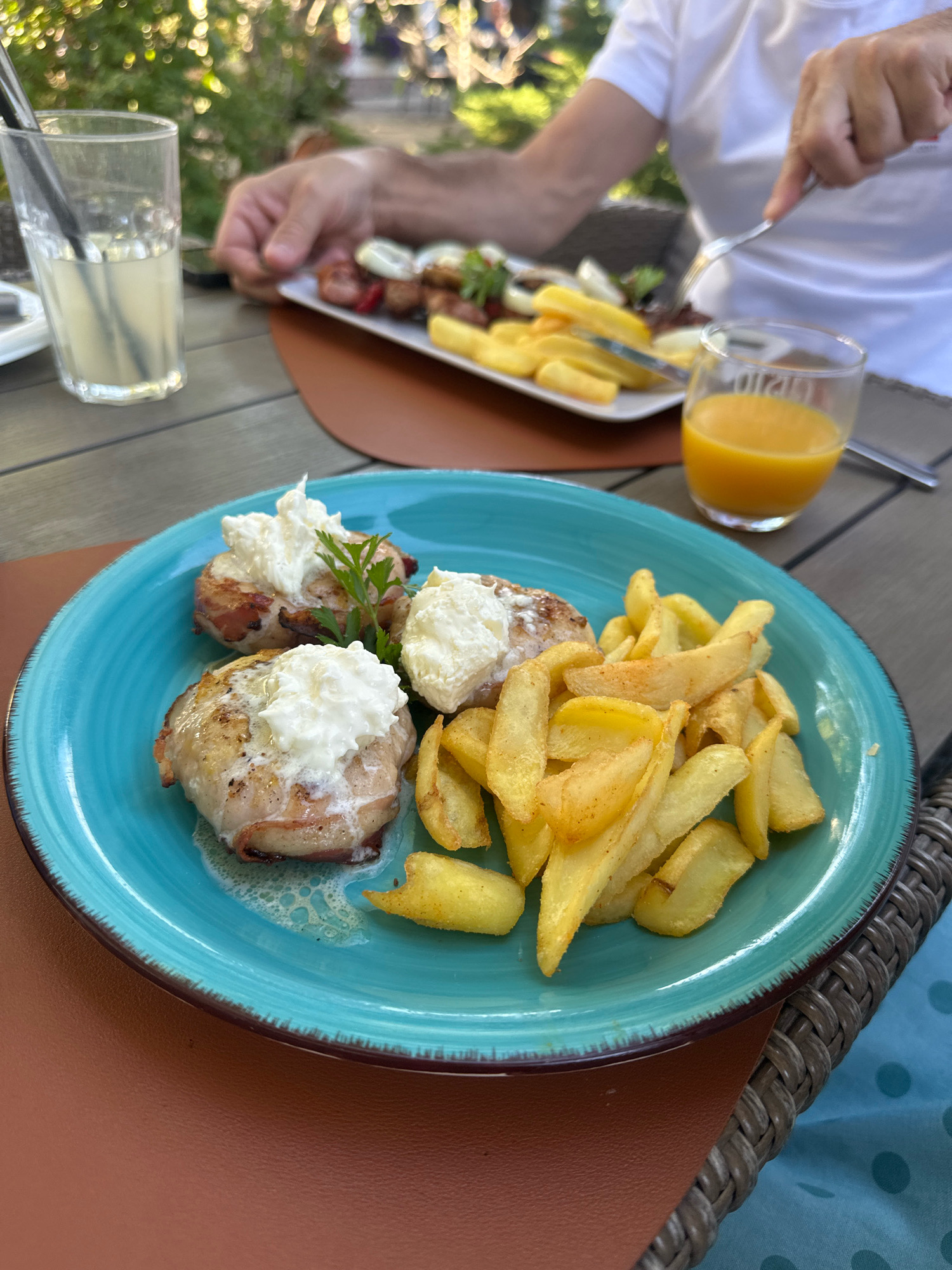In Topola Municipality, Serbia, an outdoor dining scene unfolds. A plate of three meat-wrapped dishes topped with white cheese and garnished with parsley sits beside a pile of golden french fries on a teal plate. In the background, a person in a white t-shirt uses a fork and knife to eat from another plate holding skewers of meat and vegetables with french fries. A glass of lemonade and a glass of orange juice are also visible on the wooden table. The setting appears to be a restaurant with lush green foliage in the background, suggesting a pleasant, daytime meal.