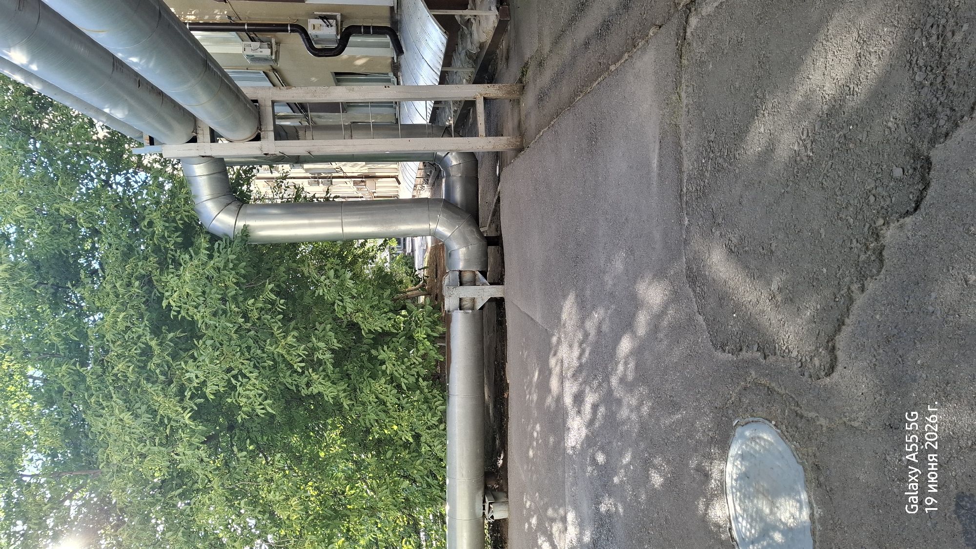 This media file captures an outdoor scene with industrial elements and lush greenery. Prominently featured are large, silver-colored pipes, likely part of a utility or industrial system, running horizontally and vertically. Some of these pipes are supported by metallic structures. Adjacent to the pipes, a dense canopy of green leaves from a tree creates a natural contrast. The ground appears to be an asphalt surface, showing signs of wear and tear, with a circular metal cover visible in the lower right portion of the image. Sunlight filters through the leaves, casting dappled shadows on the asphalt. The overall setting suggests an urban or industrial area where nature is encroaching or coexisting with man-made structures.
The time of day appears to be daytime, indicated by the bright sunlight and shadows. The weather seems clear and sunny.
Visible text in the image reads: "Galaxy A55 5G 19 июня 2026г.". This indicates the device used for capturing the image and a date in the Russian format. The location context is Rostov-on-Don, Russia.
No people or specific events are depicted. The focus is on the infrastructure and the surrounding environment.