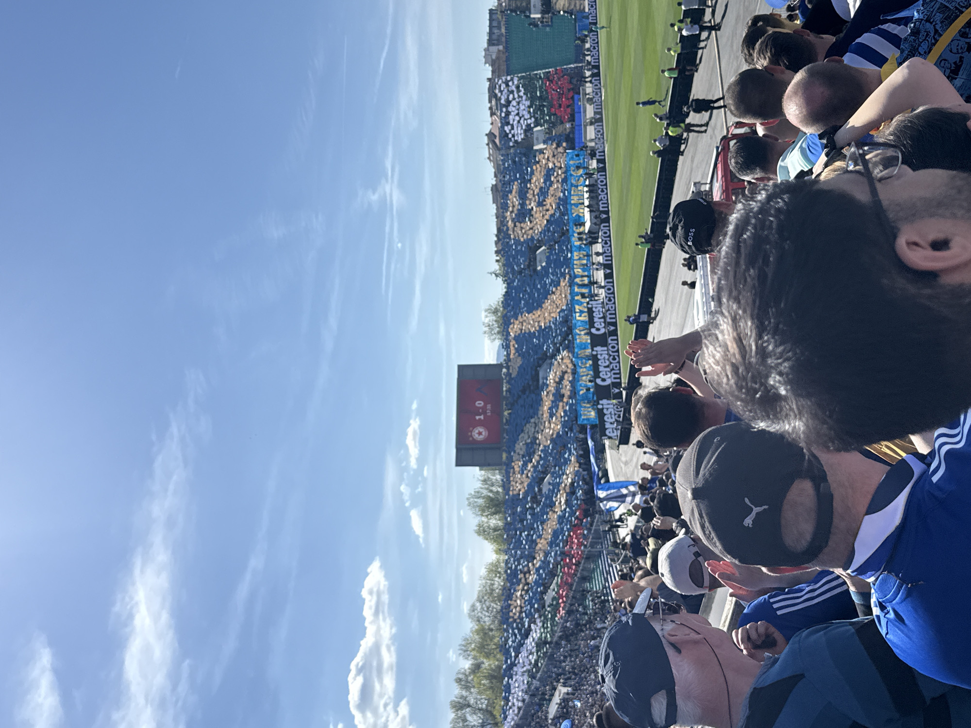 The image captures a scene at a stadium in an unspecified city, Unknown, Unknown, filled with spectators, predominantly wearing blue and yellow attire, likely supporting a soccer team. The crowd is densely packed, with faces and bodies visible in the foreground. A few wear hats, including one with a Puma logo and another with the word "BOSS." The game is in progress, as indicated by a scoreboard in the background showing the score as "1-0" and the time as "47:31." Banners with the text "МАРЕА НА БЪЛГАРИЯ СТЕ ЖИВО" are visible among the crowd. The stadium's perimeter displays advertising for "Ceresit" and "macron." The sky is blue with scattered clouds.