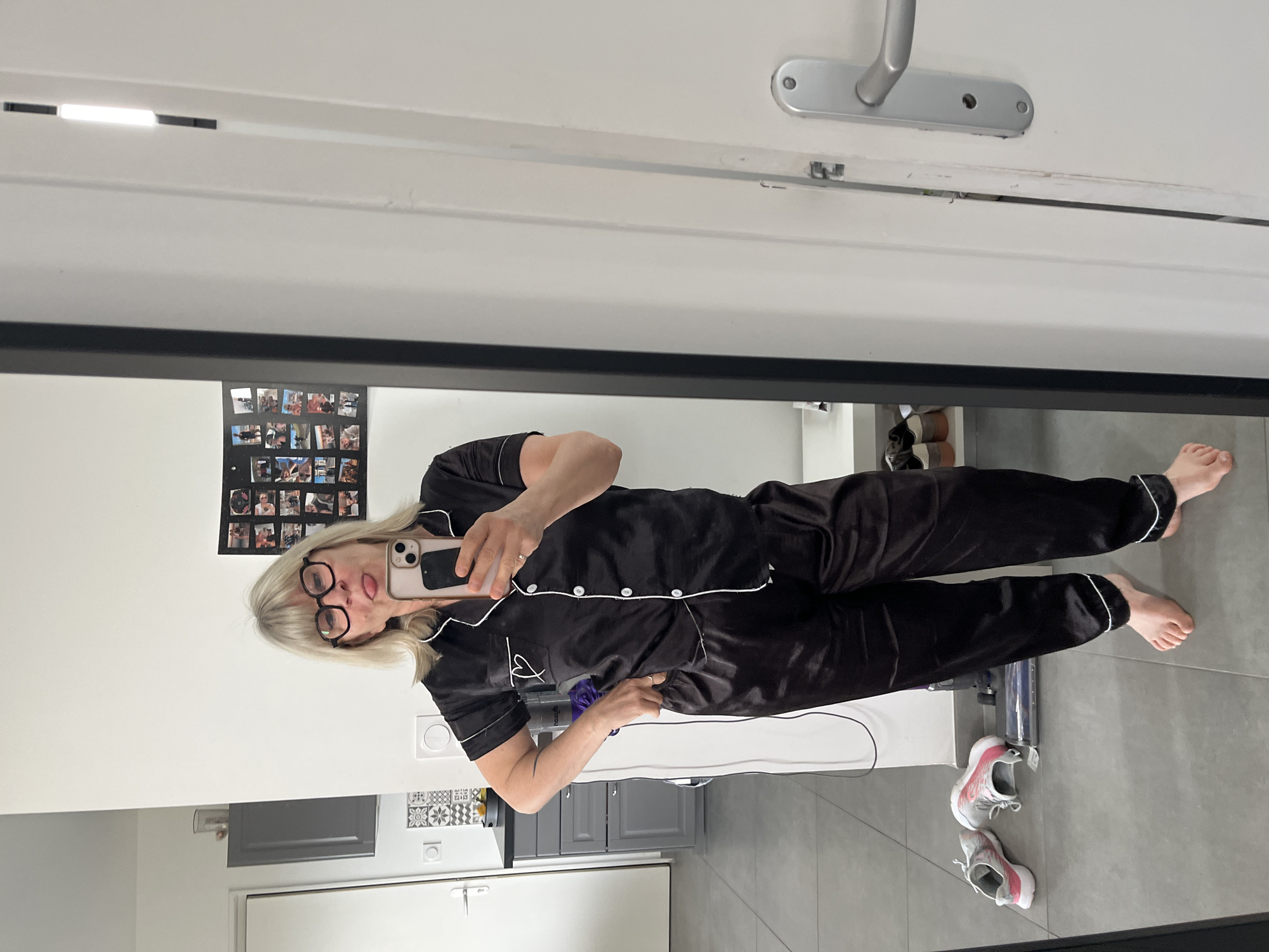 A woman with short blonde hair wearing black-rimmed glasses and black pajamas with white piping is standing in front of a mirror, taking a selfie with a smartphone. She is sticking her tongue out. The mirror reflects a room with grey cabinets, white walls, grey tile floor, and a collage of photos on the wall. A white vacuum cleaner and a pair of athletic shoes are on the floor near the woman's feet. The location is possibly in a domestic setting in Chambœuf, France, based on the context and the presence of French architectural elements.