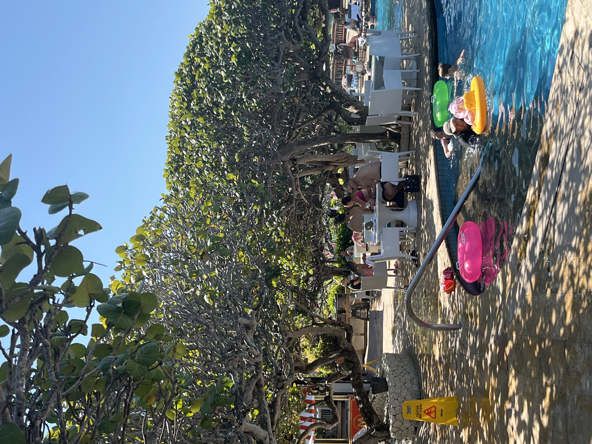 An outdoor scene in Kuta Selatan, Indonesia features a swimming pool area with people and dining tables under the shade of large trees. In the foreground, a bright pink inflatable ring floats in the blue water of the pool. To the right of the pool, a group of people are swimming, with some using inflatable rings. One person is pushing a smaller inflatable boat with a child on it. In the mid-ground, several white tables and chairs are set up under the trees, with people seated at them, seemingly dining. A yellow "caution wet floor" sign is visible on the tiled ground to the left. The background shows more trees and a clear blue sky.