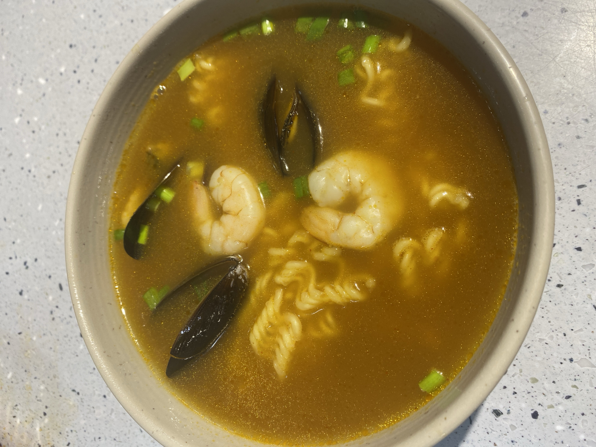 A close-up overhead shot shows a bowl of seafood ramen. The bowl is light beige with a speckled grey countertop. The ramen has a golden-brown broth, curly noodles, two cooked shrimp, two mussels, and chopped green onions.
