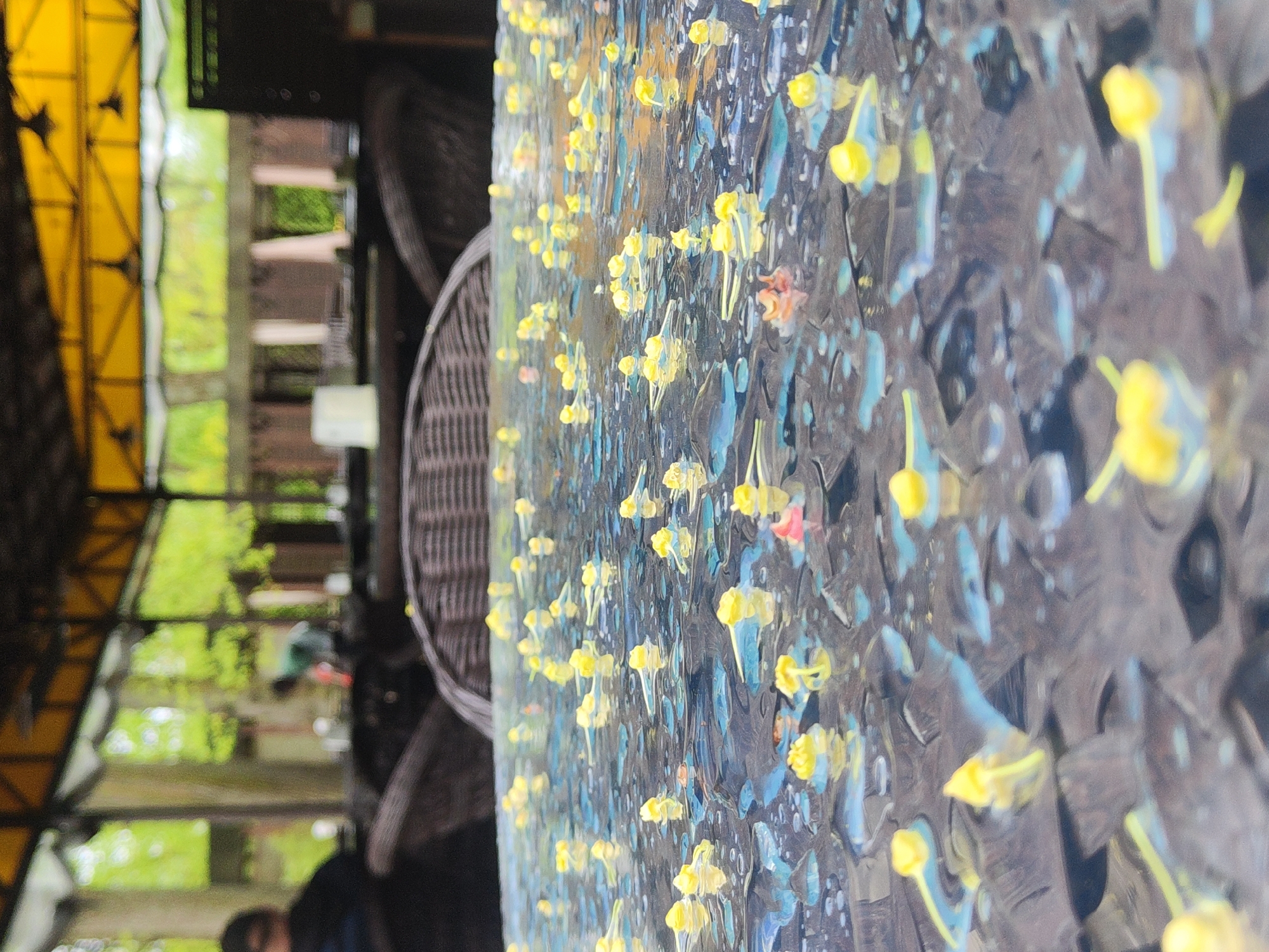 The image shows an outdoor cafe in Saint Petersburg, Russia. The cafe is covered with a yellow awning and the image is focused on a table covered in a pattern of blue and yellow flowers. There are wicker chairs and tables outside. The view behind the cafe shows trees, buildings and other people.