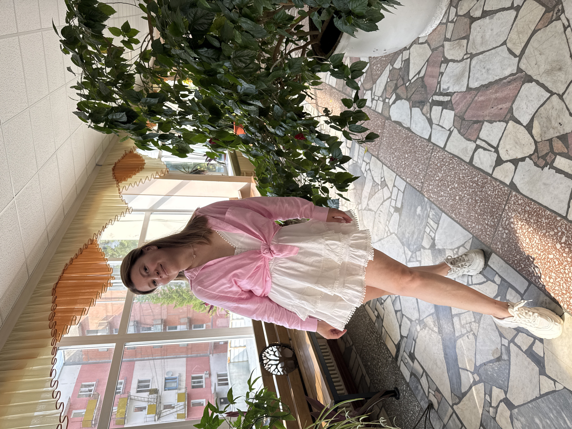 The image captures a woman posing indoors, with a large window offering a view of apartment buildings in Chaikovsky, Russia. She is wearing a pink unbuttoned shirt tied at the waist over a white ruffled mini-dress and white sneakers. To her left, a wooden bench with a decorative tree cutout is visible. A large potted plant with green leaves and some red flowers dominates the right side of the frame. The floor is tiled with a mosaic pattern of various stone shapes and colors. The overall lighting suggests daylight filtering in through the window, creating shadows on the floor.