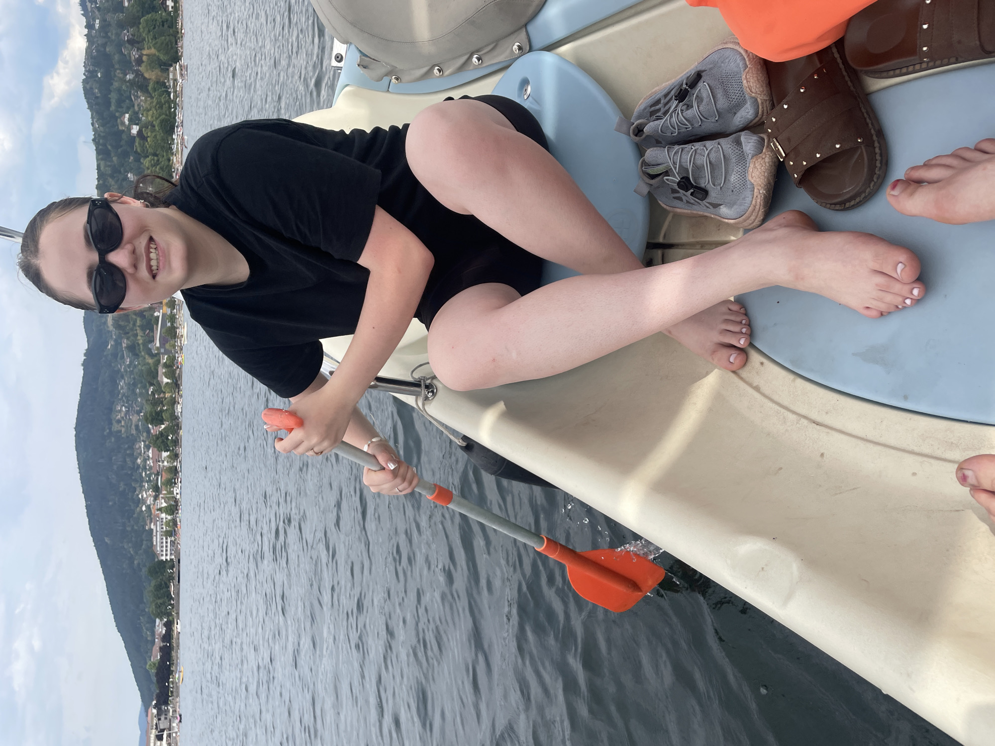 A young woman with dark hair, wearing sunglasses and a black t-shirt, is seated in a white and blue pedal boat on a lake. She is holding a paddle with an orange blade and grey shaft. Her legs are crossed, and her bare feet are visible. Next to her are a pair of grey sneakers and a brown sandal. In the background, a hillside covered in trees and buildings overlooks the lake, with several other boats on the water. This scene is taking place on Lac de Gérardmer in Gérardmer, France.