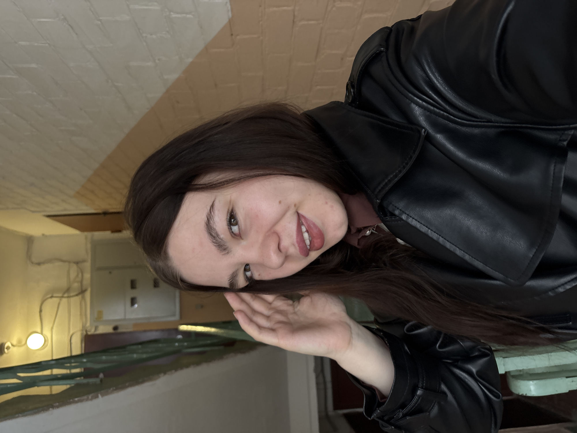 An image, correctly oriented, depicts a young woman inside an older building, likely in Perm, Russia. She has long, dark brown hair and is wearing a black leather jacket over a dark-colored top. Her head is tilted slightly, and she is smiling cheerfully at the camera with a direct gaze, her lips slightly parted to reveal teeth. Her right hand is raised to the side of her face, palm open and facing outwards, in a relaxed, possibly playful gesture. Her expressive face features prominent eyebrows and visible natural skin texture.
The background elements suggest an interior hallway or stairwell. The walls are painted in two tones: a light beige/cream and a warmer, peach-colored stripe running horizontally. Exposed electrical conduits and cables are visible along the walls and ceiling. A circular wall-mounted light fixture emits a warm, yellowish glow. Further in the background, an electrical panel or distribution box is mounted on the wall. Portions of a green or turquoise-colored railing or banister are visible in the lower left, hinting at a staircase. The overall setting appears functional and somewhat dated, characteristic of an older public or residential building. No visible text is present.