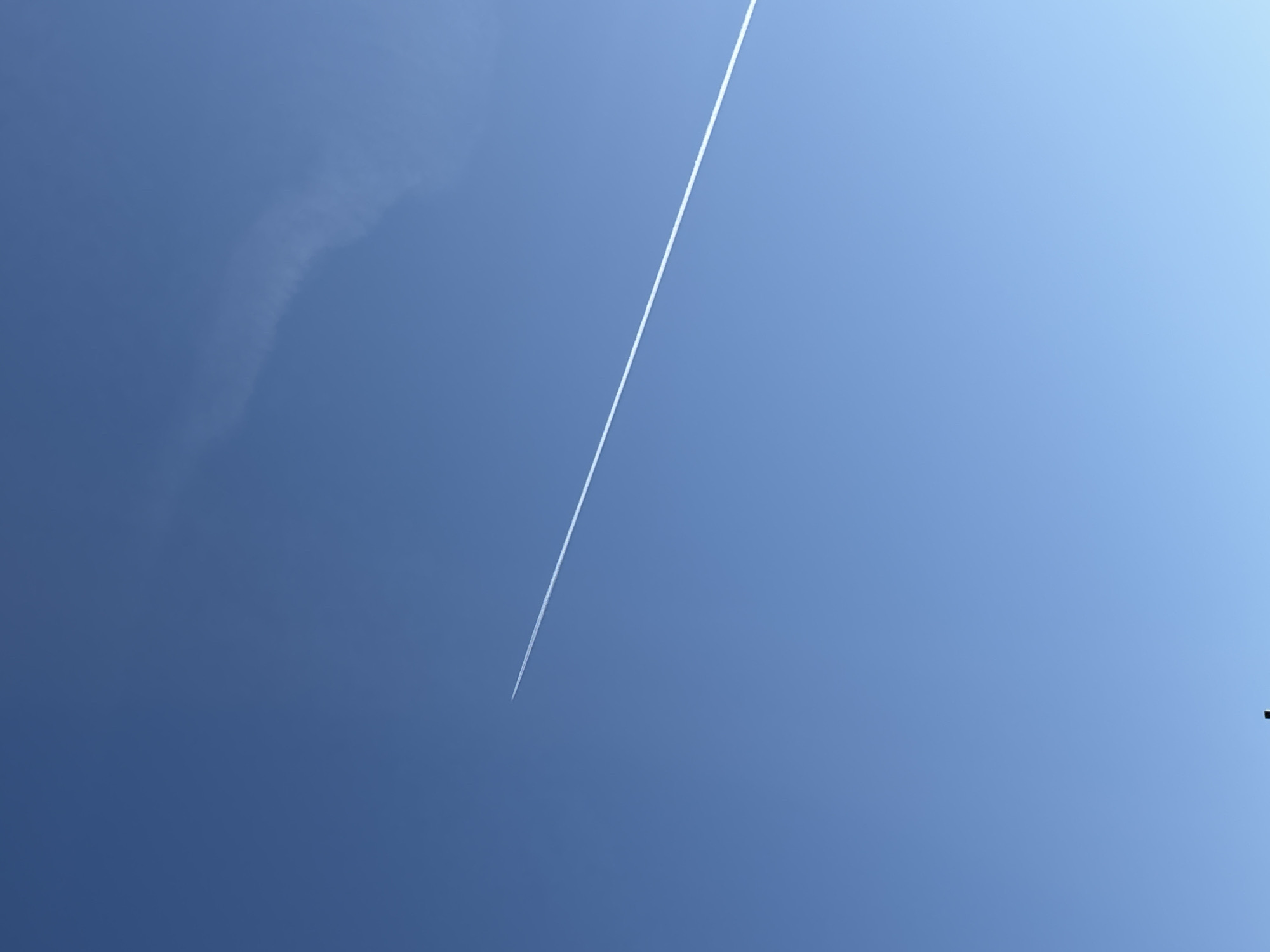 The image shows a bright blue sky with a contrail diagonally traversing the frame from the lower left to the upper center. There's a faint, blurry cloud formation in the upper left. There is no visible evidence to suggest the location is Chaikovsky, Russia. The image lacks people, objects, actions, and interactions on the ground. The environment is solely focused on the sky.