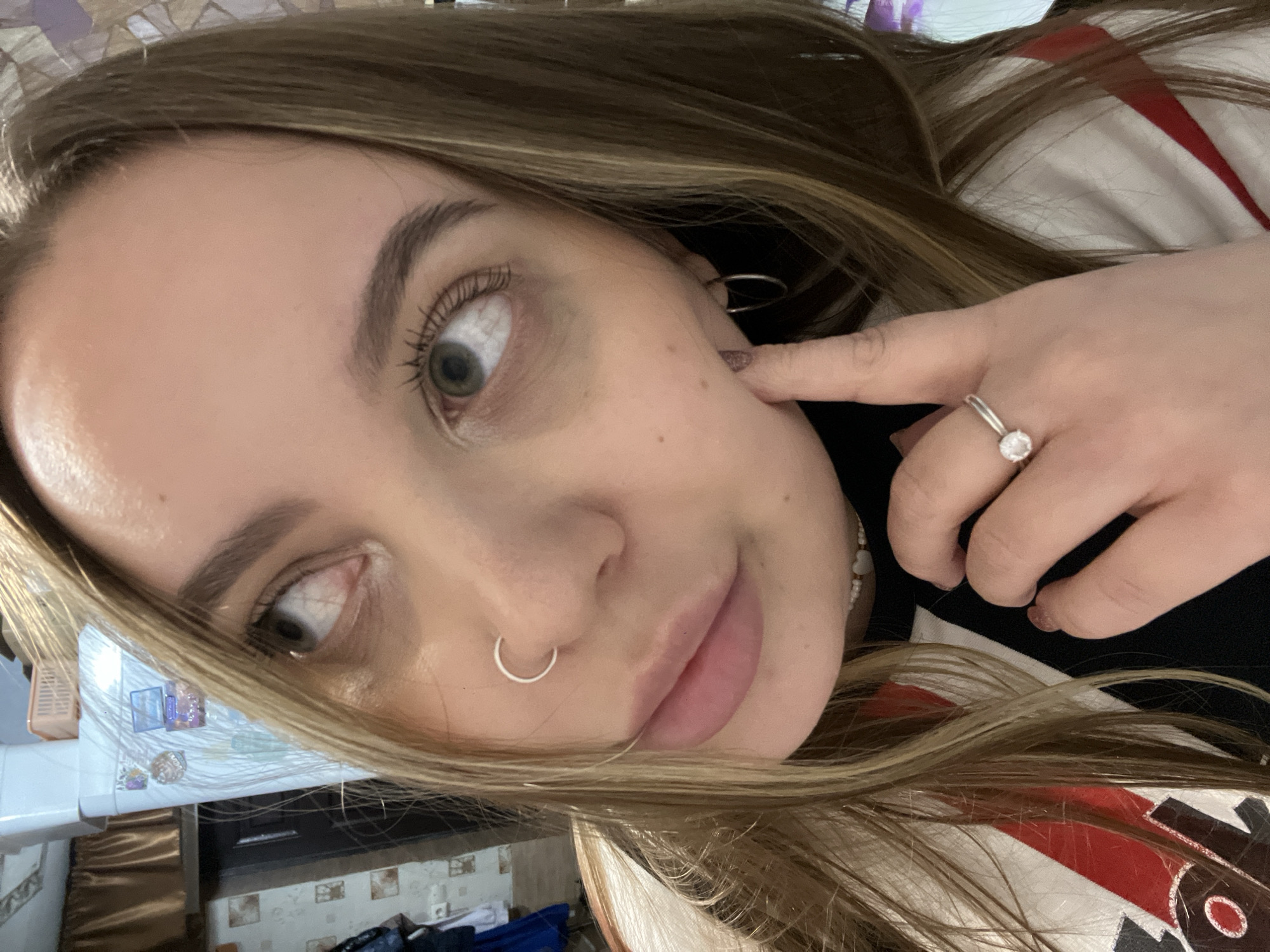 A close-up selfie of a young woman with light brown hair and green eyes. She is wearing a nose ring in her left nostril and an earring in her left ear. A ring with a prominent stone is on her left ring finger. Her left index finger is touching her left cheek. She is wearing a black shirt under a white and red patterned top. The background shows a section of a room with tiled walls, dark brown cabinets, and some personal items. The location is not discernible from the image. Novopokrovskaya, Russia, is not discernible from the image.