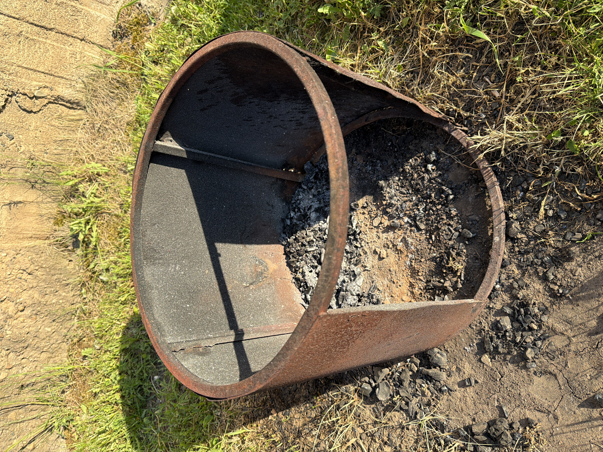 The image depicts a rusty, dilapidated metal fire pit or burn barrel, partially filled with ash and remnants of burnt material. The barrel is situated outdoors on a patch of ground consisting of bare soil, dead grass, and a few scattered stones. Adjacent to the barrel is a small section of wooden planks. The location is specified as Bolsheustyikinskoye, Russia, although no other details in the image confirm this location. There are no people or visible interactions within the frame. The primary subject is the barrel and its contents, with the surrounding environment serving as context.