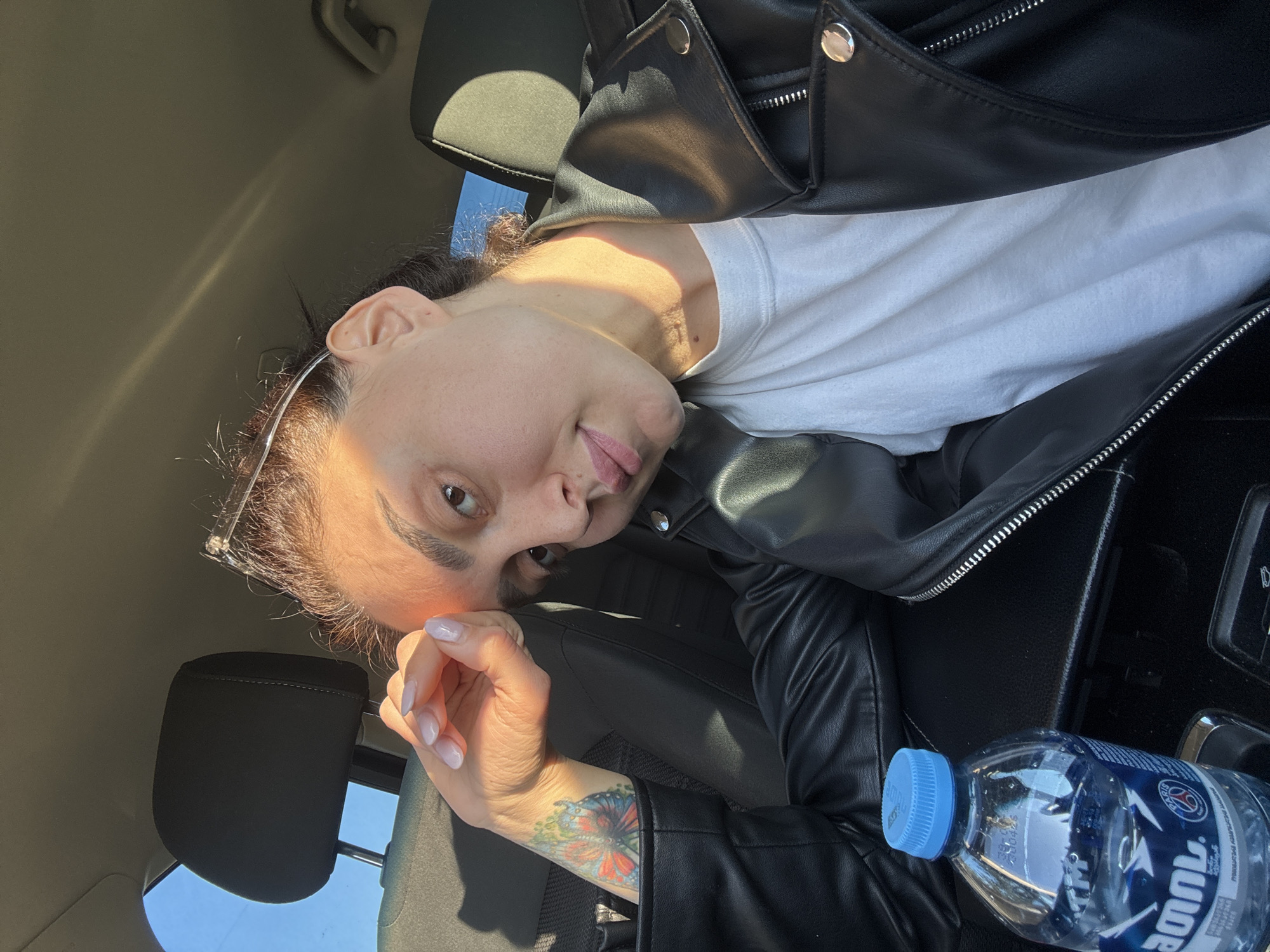 A middle-aged woman is inside a car. She wears a white t-shirt and a black leather jacket. She is looking at the camera with her head resting on her left hand. A butterfly tattoo is visible on her left arm. A water bottle is on the car's center console. The background suggests that the location is in Tbilisi, Georgia.
