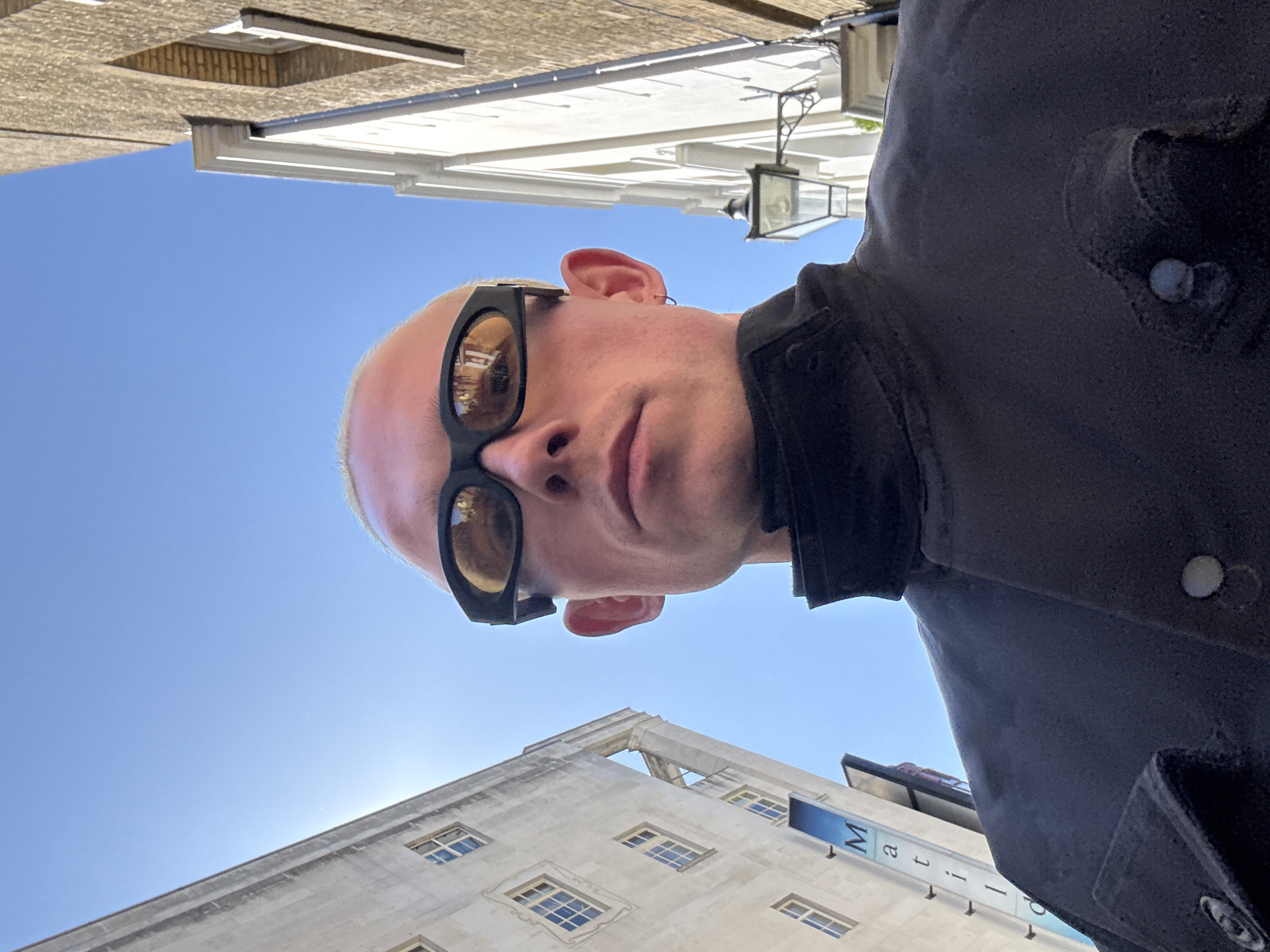 The main subject of the image is a person, likely male, with a light-colored, short-shaved or bald head, wearing oversized dark sunglasses with reflective brown lenses and a dark, possibly black, collared jacket. He is captured from a low-angle perspective, looking directly into the camera. His head is significantly tilted, rotated approximately 90 degrees clockwise relative to the background. A small earring is visible in his left ear.
The scene depicts an urban outdoor setting under a clear, bright blue sky, indicating a sunny daytime. The background consists of several buildings viewed from below. To the left side of the unrotated image (the top of the rotated image) is a large, light-colored stone or concrete building featuring numerous rectangular windows, some with multiple panes. A blue banner with white text is affixed to this building, where partial letters "M a r i" and "l l e r" are visible. To the right side of the unrotated image (the bottom of the rotated image), other buildings are partially seen, including one with a textured, possibly brick or stone facade, and another lighter-colored building adorned with traditional-style ornate street lamps and brackets. The bright sunlight creates reflections in the sunglasses and a slight lens flare in the sky. The activity appears to be a casual portrait or selfie taken in a city environment, likely London, United Kingdom, given the architectural style.