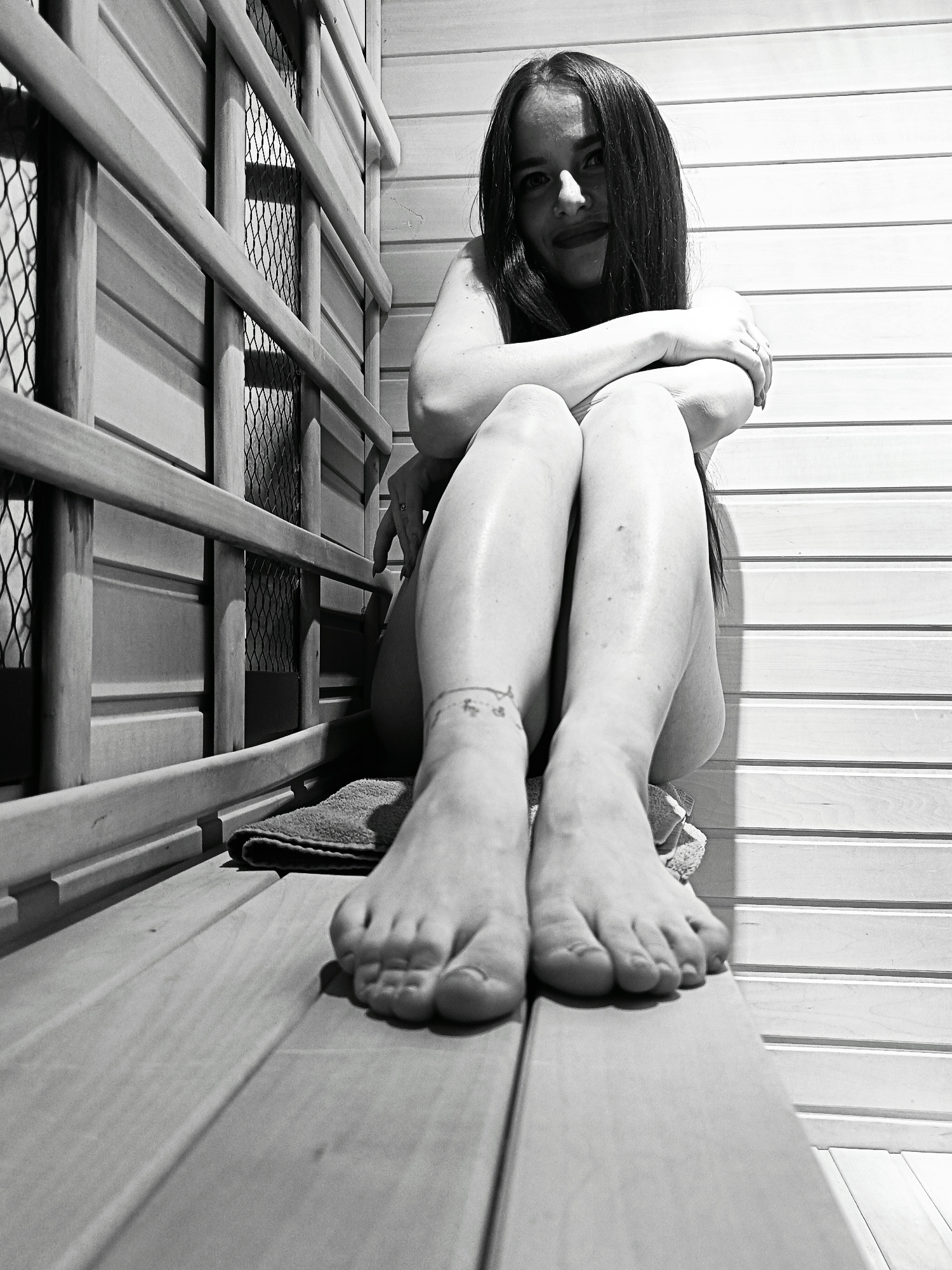 A black and white image captures a young woman seated within what appears to be the interior of a sauna or a wood-paneled room. She is positioned at a low angle, gazing directly into the camera with a slight smile. Her long, dark hair frames her face. Her arms are crossed over her chest, and her legs are bent with her feet prominent in the foreground, pointing towards the viewer.
The setting is defined by light-colored wooden slats and benches. To the left, a vertical structure featuring wooden bars and a metal mesh grating suggests a protective cover for a heat source or lighting. The wall to the right consists of horizontal wooden planks.
The woman appears to be bare-chested or minimally clothed, with her skin exhibiting a subtle sheen, possibly from moisture or sweat. A delicate anklet, adorned with small, indistinct charms or symbols, is visible on her left ankle. A ring is noticeable on her left ring finger. A folded towel rests on the wooden bench beside her. The strong lighting creates distinct contrasts and shadows throughout the scene, emphasizing the textures of the wood and the woman's features. No discernible text is visible in the image.