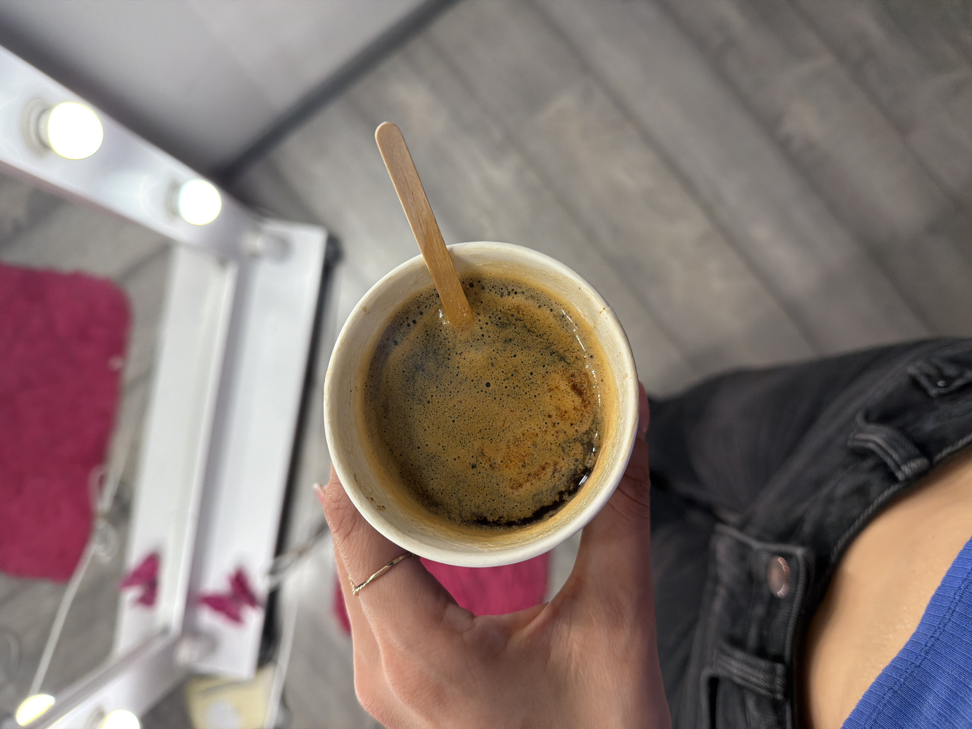 An overhead shot shows a hand holding a paper cup of coffee with a wooden stirrer. The coffee is dark brown with a foamy surface. The hand has a thin gold ring on one finger. In the background, a vanity mirror with lights is visible, with a pink fuzzy rug reflected in it. The floor appears to be made of grey wood planks. The lower part of the image shows the person's dark denim jeans and a blue garment. The scene is likely indoors, possibly in a dressing room or backstage area, given the vanity mirror. The location is not specified in the image or provided context.