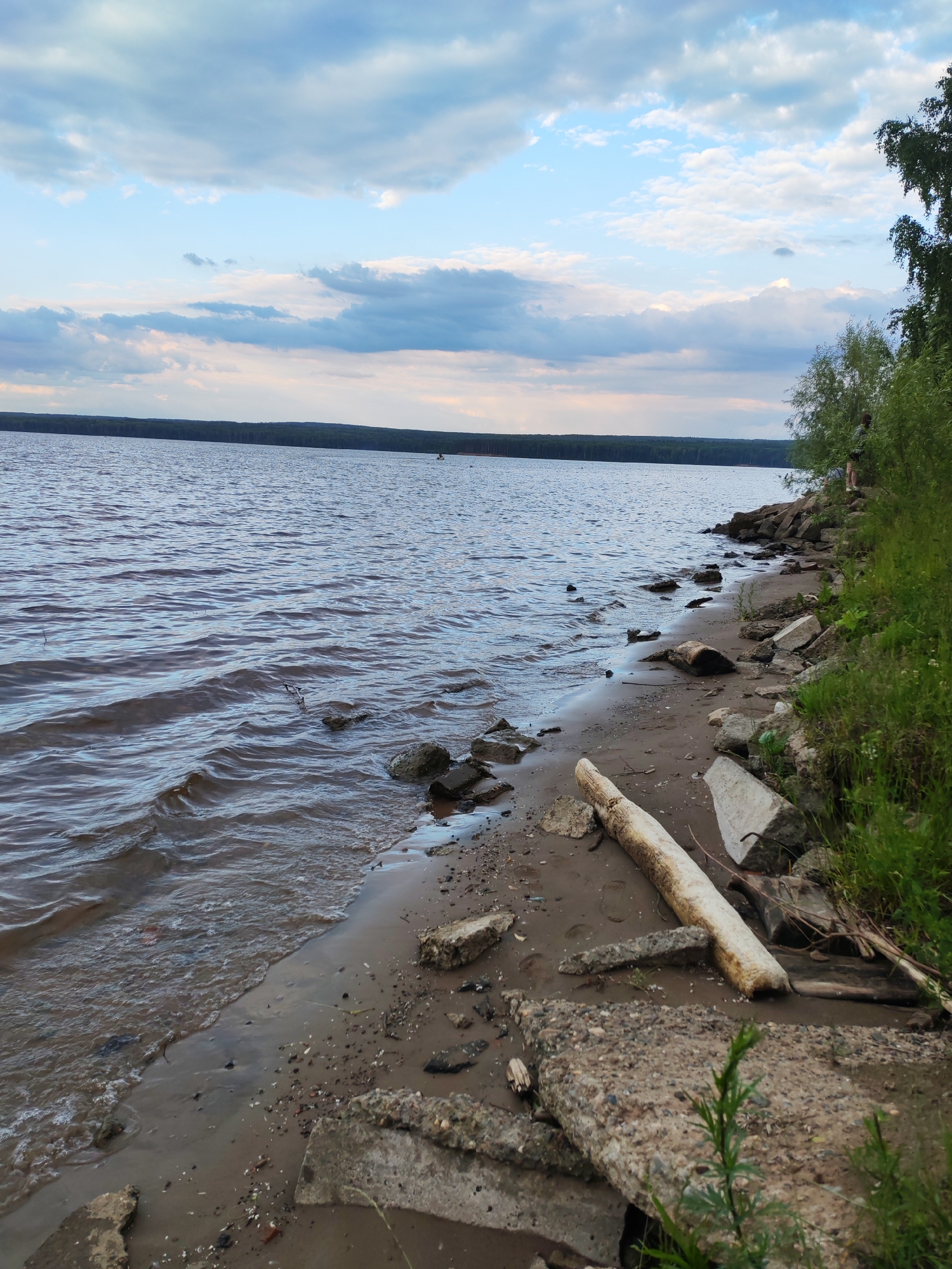 This is a landscape photo of a body of water with a sandy and rocky shore. The water is calm with gentle ripples and appears to be a lake or a wide river. In the distance, a dark line of trees marks the opposite shore. The sky is overcast with a mix of white and gray clouds, suggesting it is either morning or late afternoon.
The foreground of the image shows a shoreline cluttered with various objects, including rocks, driftwood, and fragments of concrete. The sand is damp from the water's edge, and some patches of grass and weeds are growing among the debris. To the right of the frame, lush green foliage and trees form a dense embankment.
There are a few subtle hints of human presence. Far in the distance on the water, a small dark shape might be a boat or a person. On the grassy embankment, two figures are partially visible, appearing to be standing or sitting. Their presence adds a sense of tranquility to the scene, suggesting a peaceful outing by the water.
The overall mood of the image is serene and natural, with a slight hint of human impact through the scattered debris on the shore. The lighting suggests a soft, diffused natural light, typical of cloudy weather. The location context of Chaikovsky, Russia, indicates a possibly rural or semi-rural setting with access to a significant body of water. No text is visible in the image.