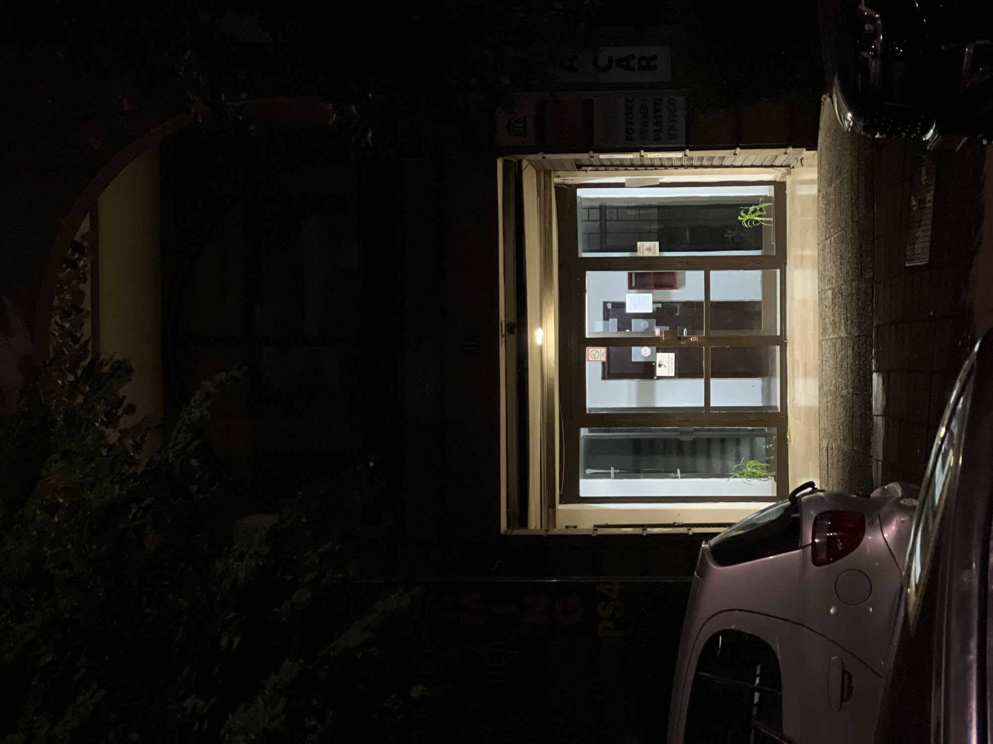 The image depicts the entrance of a building at night. The entrance features double glass doors, illuminated from within, revealing a staircase behind them. Small potted plants are visible on either side of the entrance. To the left of the entrance, partially obscured by darkness and foliage, is a dark sign with vertical text. To the right, there are several smaller signs mounted on the wall. In the foreground, a light-colored car, possibly a hatchback, is parked. The scene appears to be in a city setting, with the presence of cars and building structures. The location is identified as Belgrade, Serbia. The overall atmosphere is dark and quiet, suggesting a time outside of typical business hours. There are no people visible in the image.