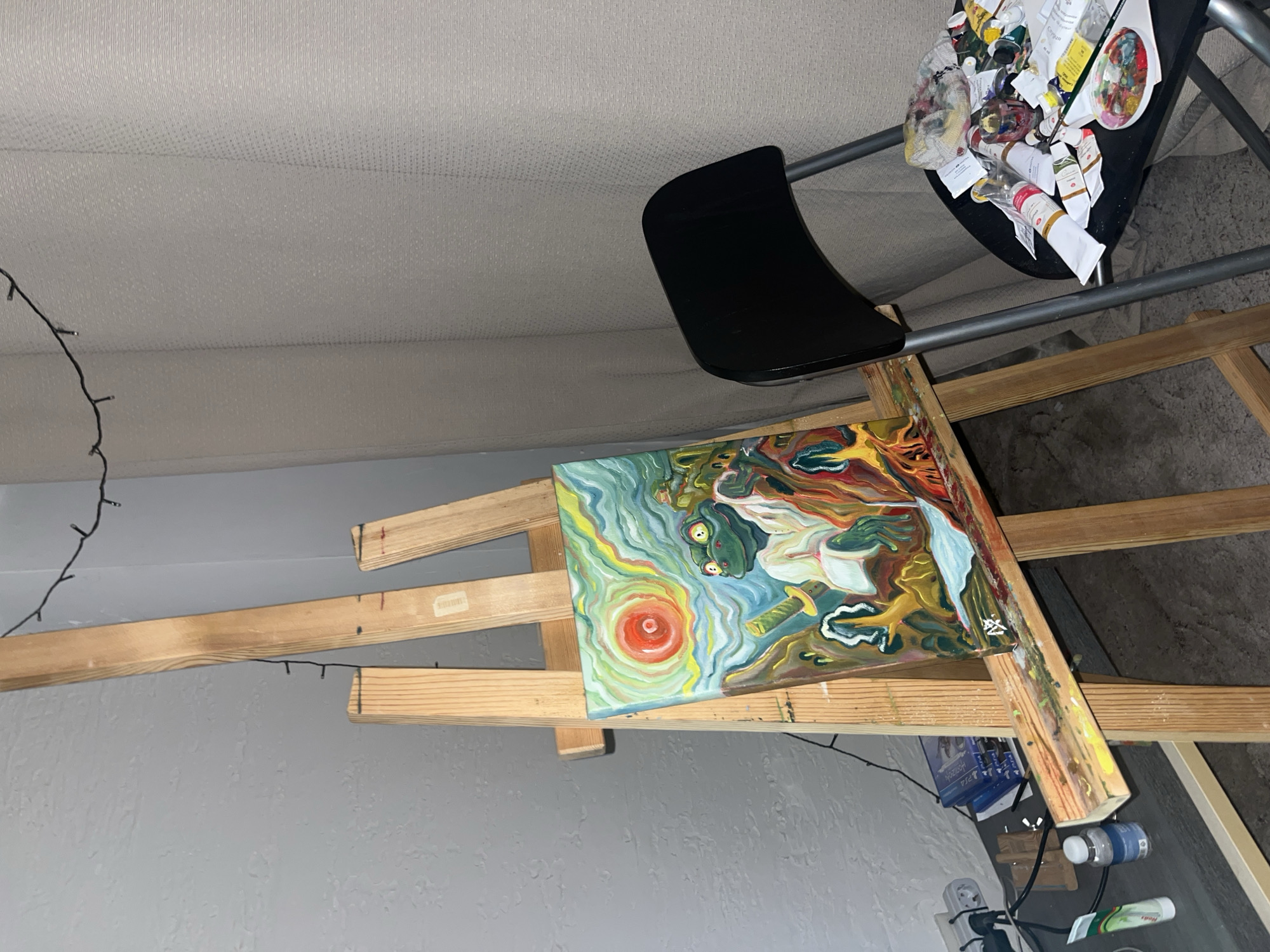 A still-life scene shows an unfinished painting on a wooden easel. The painting depicts an abstract landscape with a prominent red sun, light blue sky, and other colors. The easel stands against a grey wall. A string of lights hangs near the wall. A black chair is positioned beside the easel, cluttered with paint tubes and painting supplies. The location of this image is not able to be determined to be Krasnoyarsk, Russia.