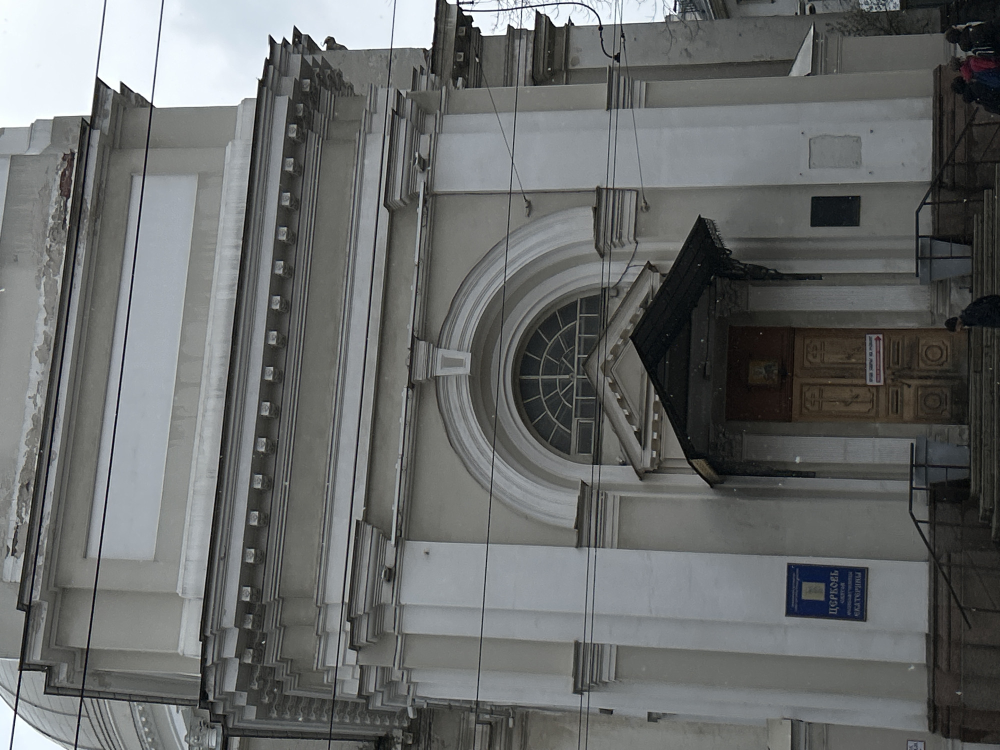 The image shows a portion of the facade of the Church of St. Catherine on Vasilevsky Island in Saint Petersburg, Russia. The building features a light-colored facade with multiple levels of architectural detailing, including stepped sections and decorative moldings. A prominent circular window is visible, along with a dark awning over the entrance. A blue sign in Cyrillic script is fixed to the wall beside the entryway. Some individuals are visible near the entrance, and there are numerous utility wires in the foreground. The weather appears overcast.