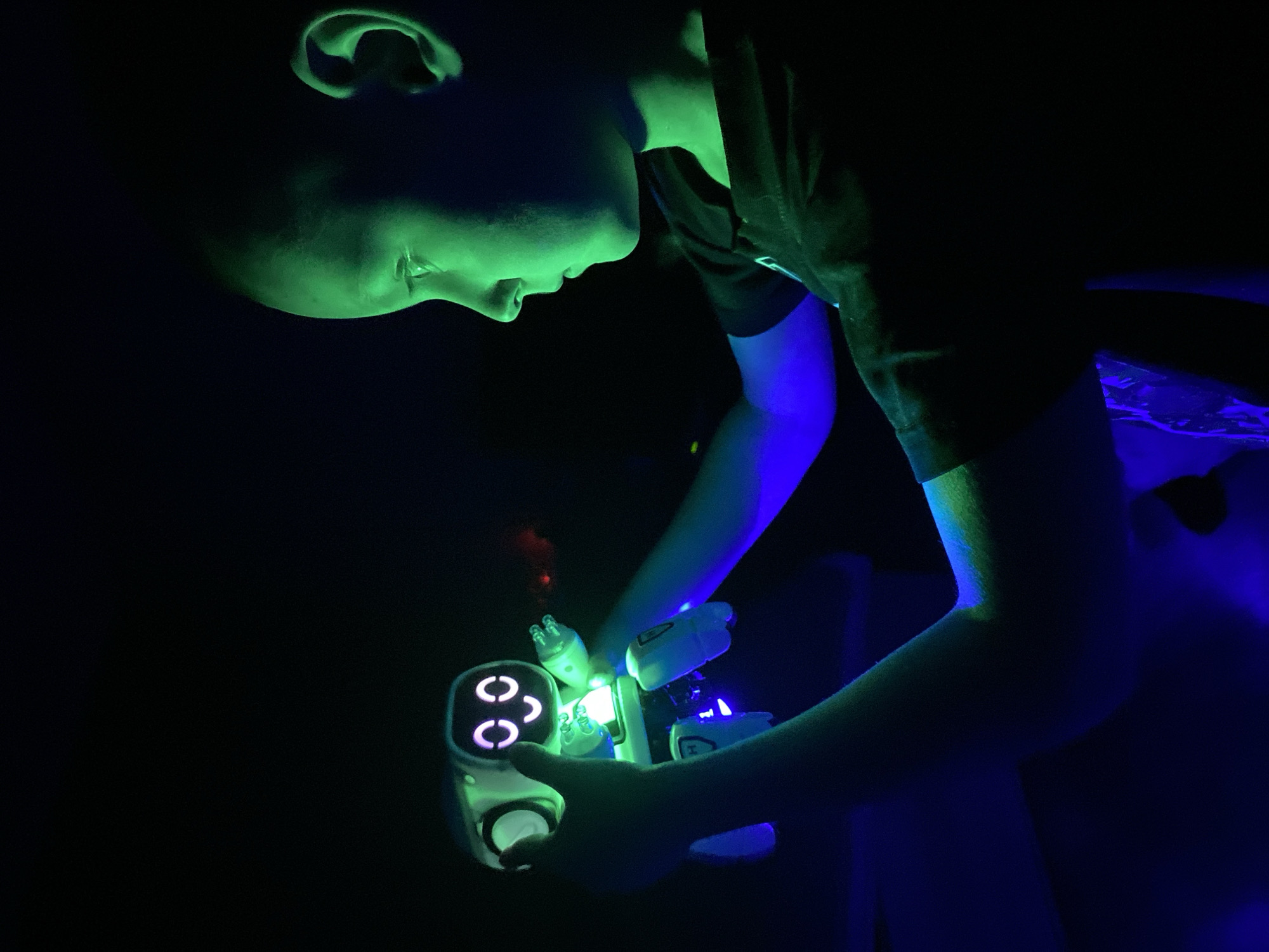 A young person, likely a child or adolescent, is depicted in a dark, low-light environment, possibly indoors at night. Their profile, including their face, ear, and parts of their upper body and arms, is illuminated by vibrant, colored light. The light casting upon their face and ear is a distinct green, while their arms and parts of their clothing are bathed in blue and purple hues.
The central activity involves the person intently holding and interacting with a glowing white-bodied device. This gadget, possibly a toy or educational tool, emits its own bright light. Notable features of the device include three glowing, pink or purple 'C' shaped symbols arranged vertically on its main body. Additionally, several smaller, cylindrical attachments extend from the device, each emitting a white or green light, with one showing a faint black letter 'H'.
The overall scene is characterized by dramatic contrast between the intense, localized colored illumination and the almost entirely black background, providing no specific location cues for Noviy, Russia, or the time of day beyond a dark setting. The person's expression appears focused and engaged with the glowing object.