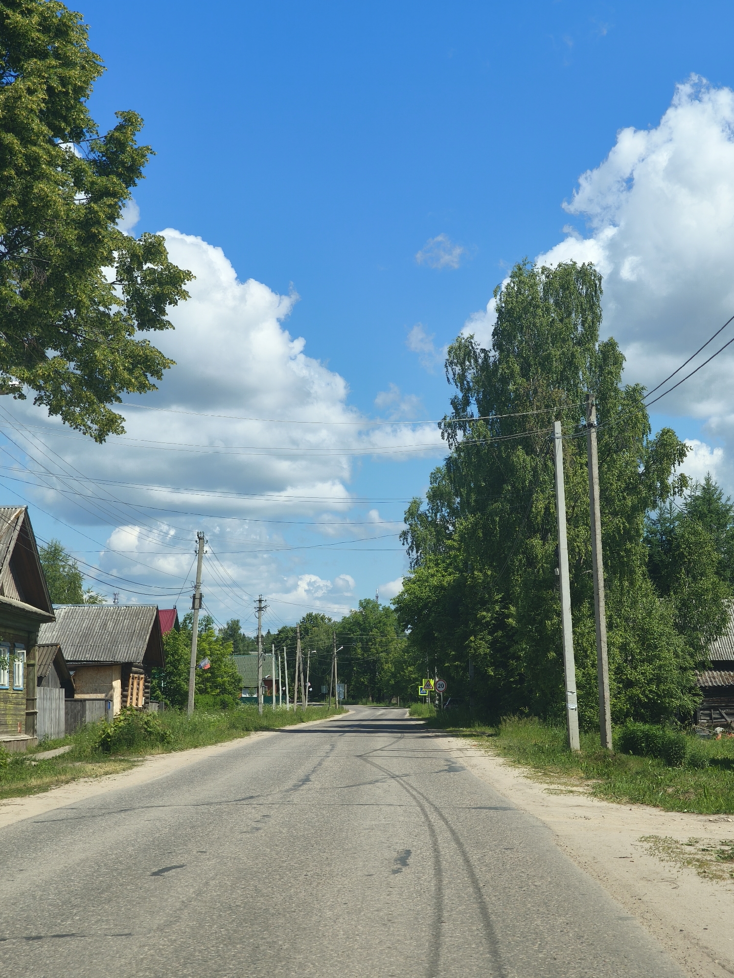 The image depicts a rural road in Kady, Russia, on a sunny day with a bright blue sky dotted with white clouds. The road, appearing to be asphalt, curves gently to the right and shows signs of tire tracks. On the left side of the road, a wooden house with green siding and white-trimmed windows is visible, along with other older wooden structures with corrugated roofs. A large, leafy tree extends into the upper left corner of the frame. On the right side of the road, a tall birch tree dominates the scene, with several electrical poles and overhead wires running parallel to the road. Dense green foliage lines both sides of the road, suggesting a village or countryside setting. In the distance, more houses and trees are visible, with a yellow pedestrian crossing sign and a circular speed limit sign (indicating 20) at an intersection. The overall atmosphere is tranquil and suggests a typical summer day in a Russian rural community. There are no people visible in the image.
