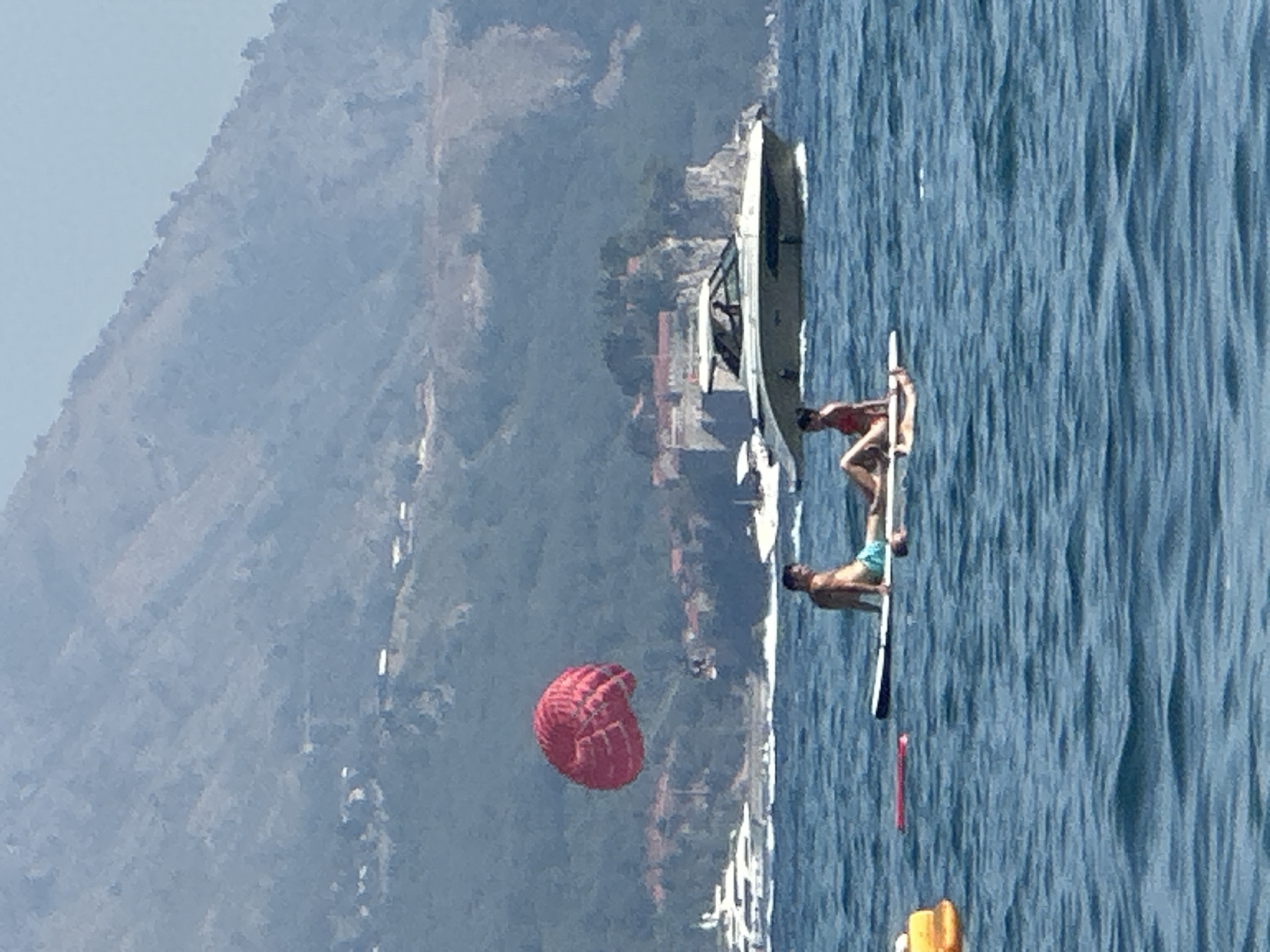 In the city of Boreti, Montenegro, two individuals are seen relaxing on a stand-up paddleboard on a body of water. The male is seated with his back to the camera, wearing blue shorts. The female is also seated, facing the camera, wearing a red bikini. The paddleboard is white with black accents. In the background, a large, red parasail floats in the sky. Several boats are visible in the distance, including a white yacht and a sleek, green and white motorboat. The backdrop consists of a lush, green, mountainous landscape with some buildings visible on the slopes. The water is a deep blue with gentle ripples. The scene appears to be a leisurely day on the water.