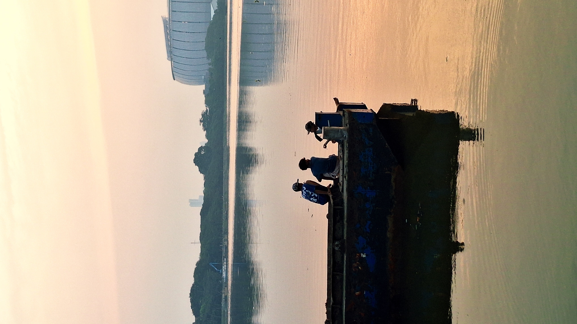 The image depicts a serene body of water at sunset, likely Ancol Bay in North Jakarta, Indonesia, given the presence of a large, modern stadium in the background which resembles the Gelora Bung Karno Stadium. In the foreground, three young individuals are seated on the edge of a dark, weathered barge. The person on the left wears a blue cap and a blue t-shirt with white lettering, facing away from the camera. The person in the middle, also facing away, is dressed in a blue t-shirt and white shorts. The individual on the right, facing the camera and wearing a cap, appears to be holding something in their hands, possibly a fishing rod. The water reflects the warm hues of the sunset, casting an orange glow on the scene. Lush green trees line the distant shore, and a few modern buildings, including the prominent stadium, are visible against the hazy sky. The overall atmosphere is one of peaceful contemplation and leisure as the day draws to a close.