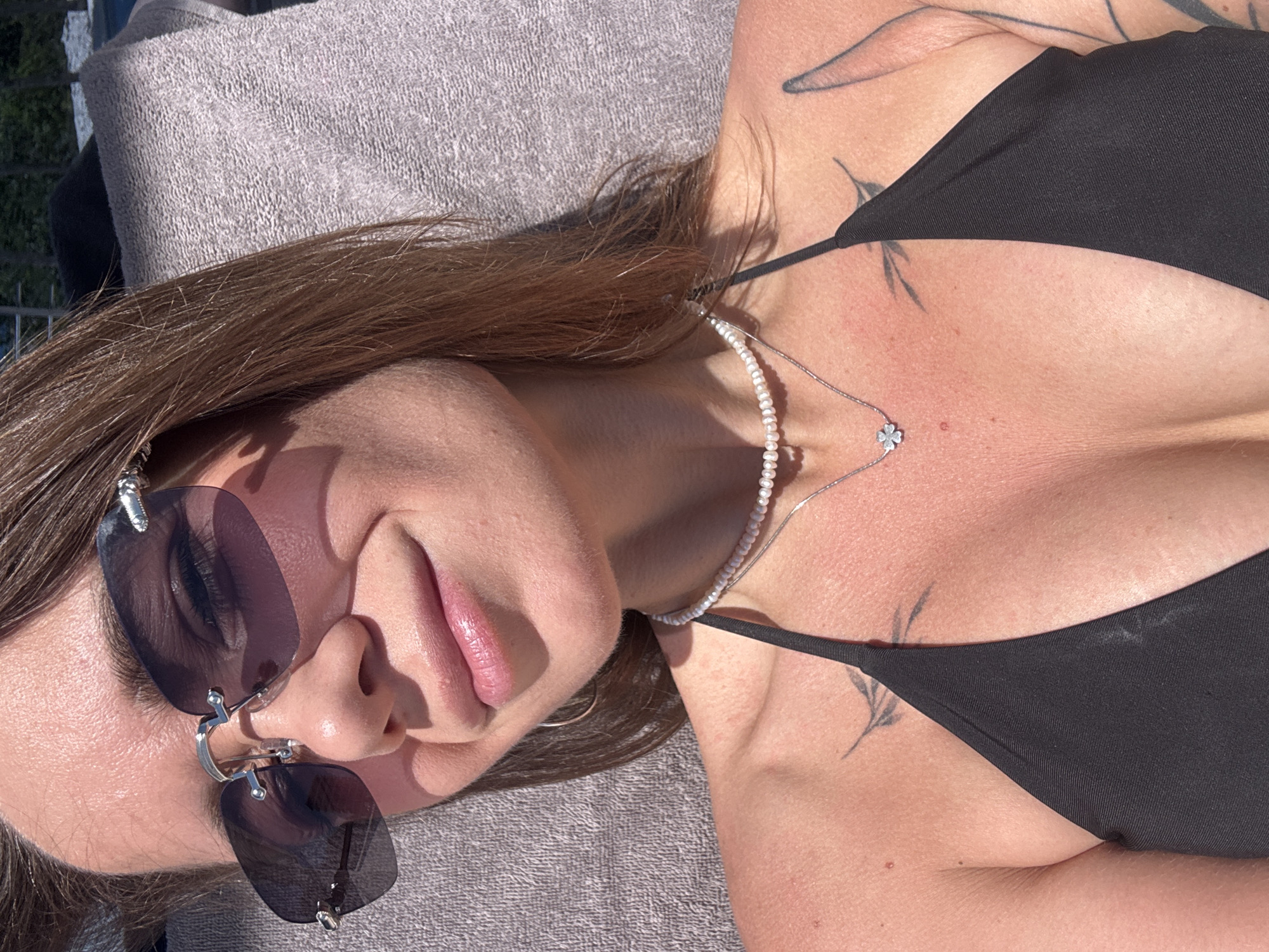 A woman with brown hair, wearing sunglasses and a black bikini top, is lying on a gray towel. She is wearing a necklace with a pearl strand and a clover-shaped pendant. She has tattoos on her chest and neck. The location is not specified as the city of Boreti, Montenegro.