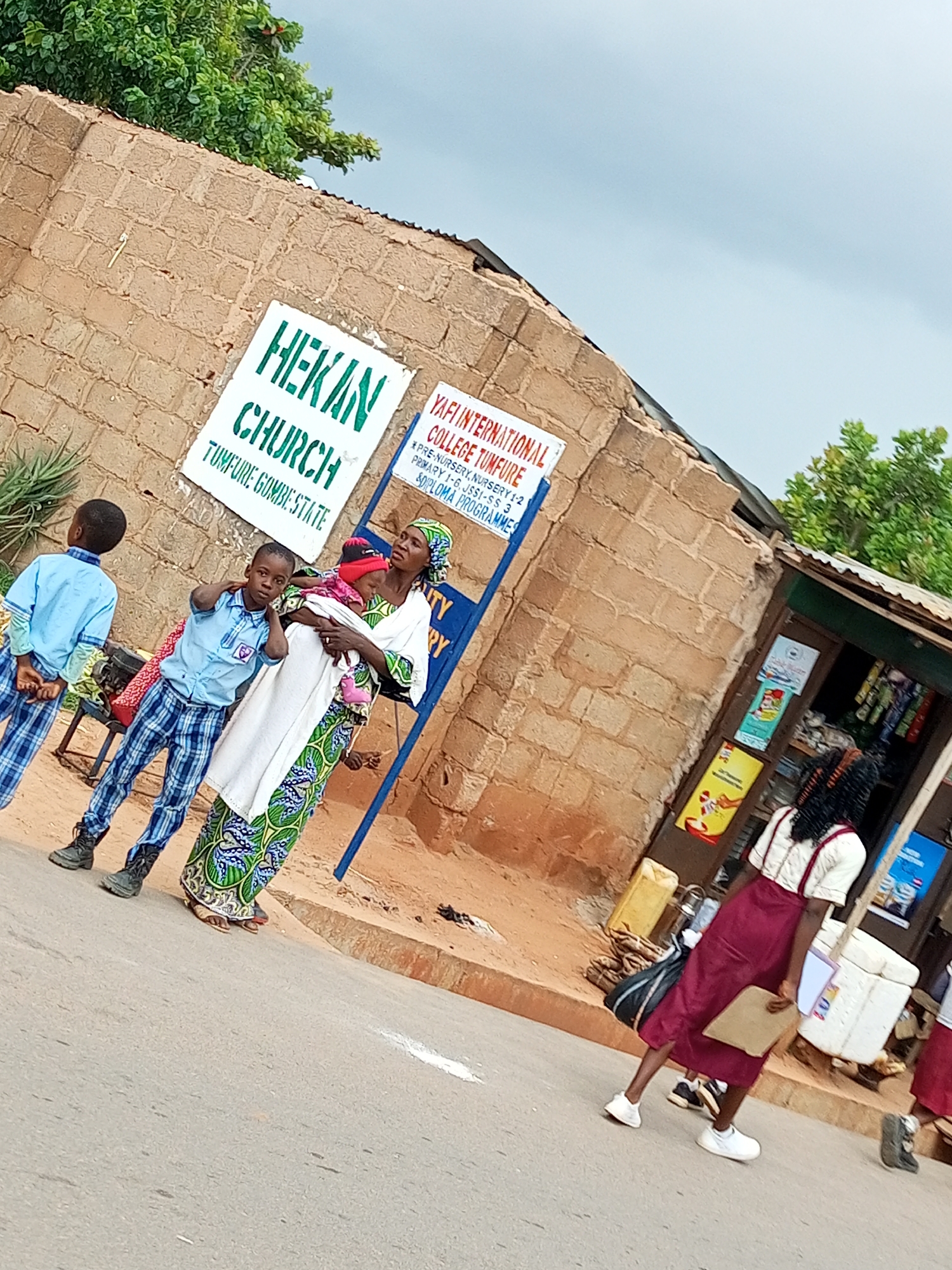 A group of people are gathered outside a brick building in Tempure, Nigeria. Two young boys, one in a light blue shirt and plaid pants, the other in a blue shirt and plaid pants, stand on the left side of the image. A woman holding a baby is in the center, wearing a white top and a colorful patterned skirt and headscarf. To the right, a young woman in a maroon skirt and white shirt with suspenders walks away from the camera. Behind them, a storefront displays various items. Two signs are visible on the building: one reads "HEKAN CHURCH TOMPURE GOMBE STATE" in green lettering, and another, partially obscured, advertises "YAFI INTERNATIONAL COLLEGE TUMFIRE." The sky is cloudy.