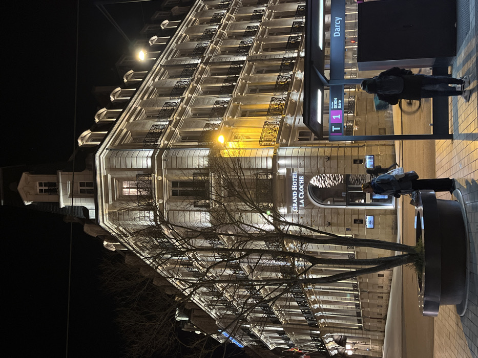 A tall, well-lit building, the Grand Hotel La Cloche, dominates the nighttime view. Bare tree branches reach up in front of it. A bus stop is visible, with "Darcy" and "T 1" markings. Pedestrians wait at the stop and near a circular planter. Streetlights illuminate the scene. The image is oriented sideways.