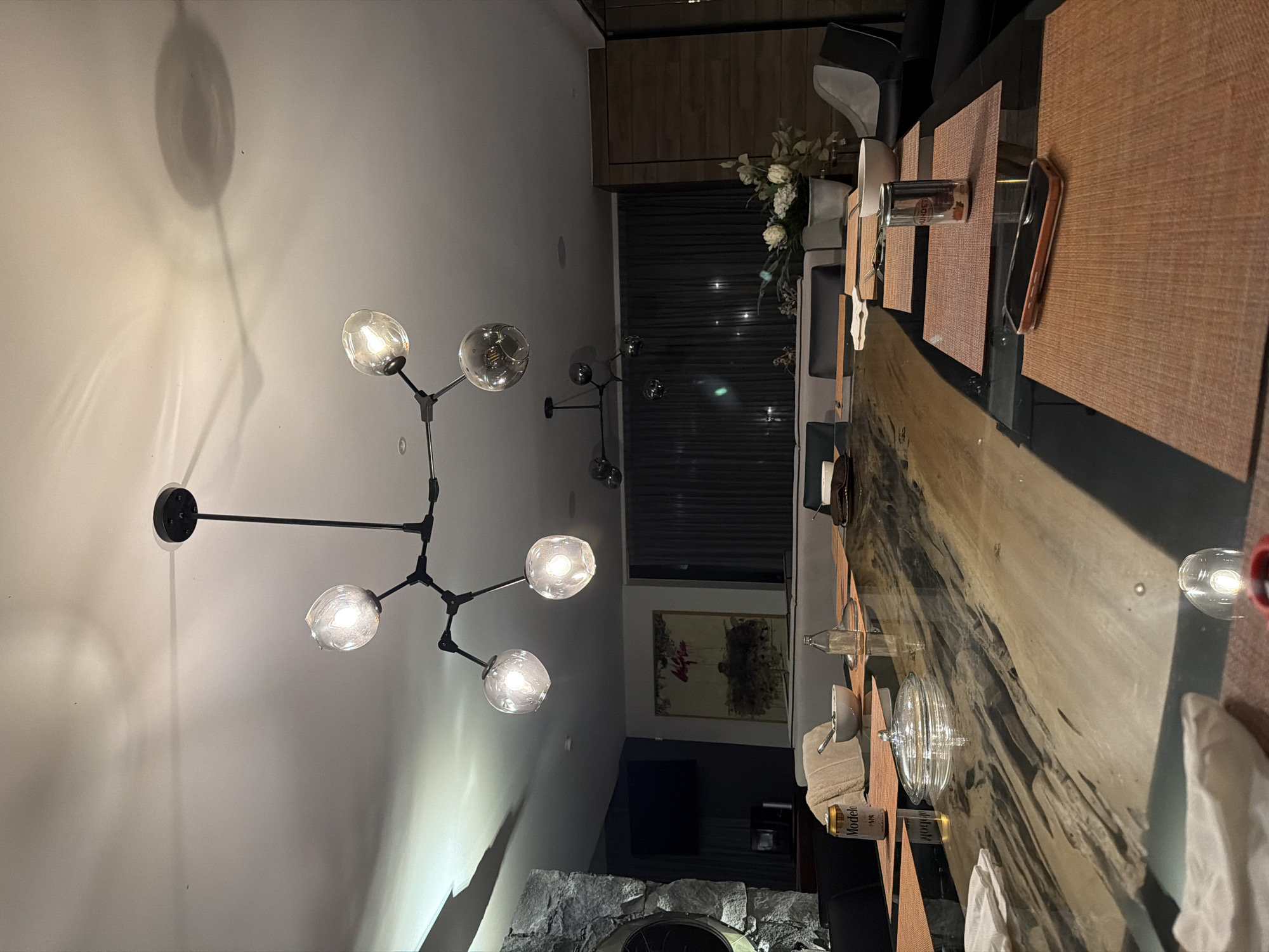 The image depicts an interior dining space. A long wooden table, set with place mats, plates, glasses, and napkins, dominates the foreground. A modern chandelier with multiple globe lights hangs above the table. In the background, there are window blinds, a cabinet, a piece of art, a television, and a stone feature. The scene is lit with artificial light. The location is Santa Ana Yancuitlalpan (Chilhuacán), Mexico. There are no people visible.