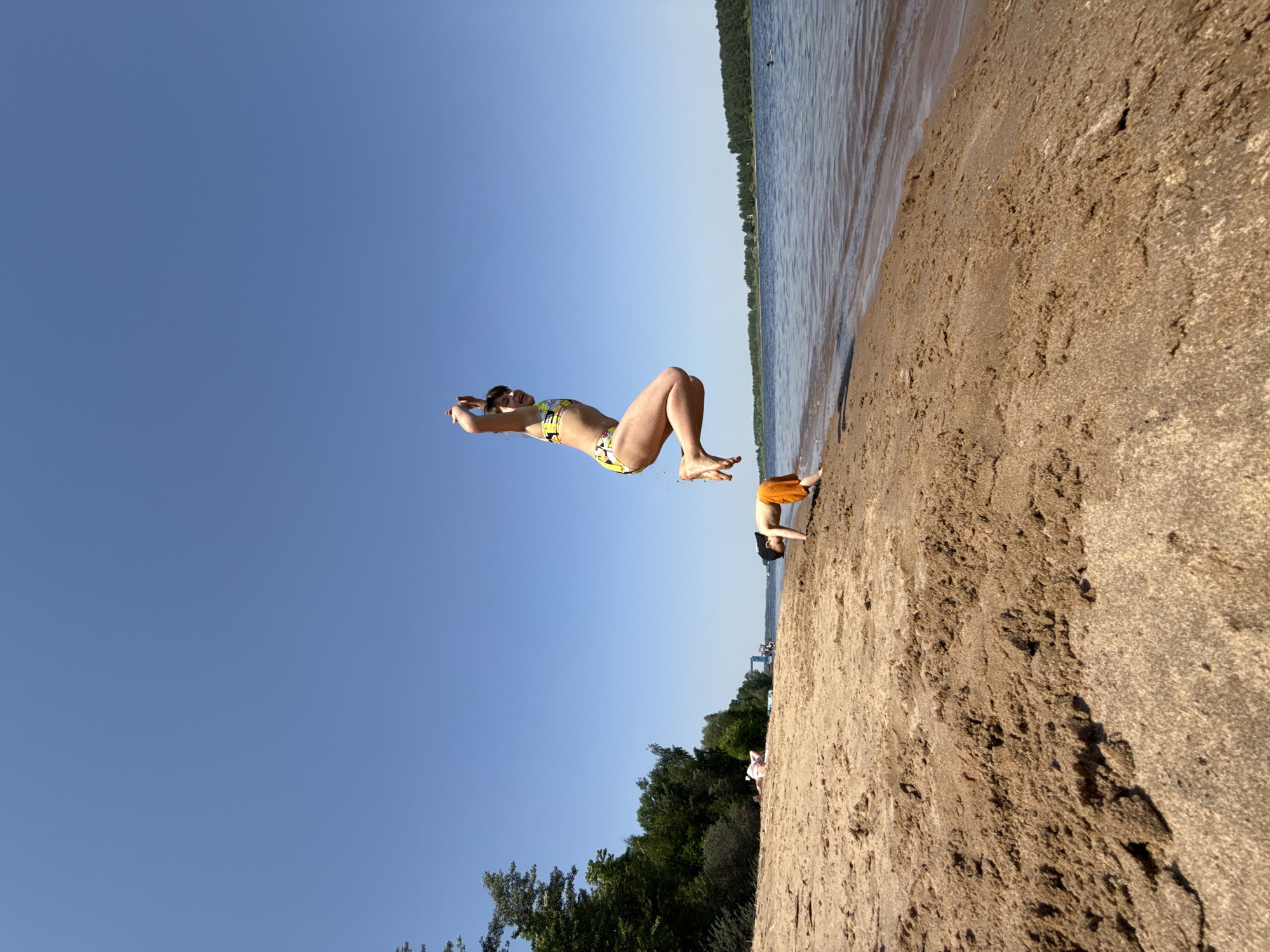 The photograph shows a woman in mid-air, seemingly performing a backflip on a beach. She is wearing a patterned bikini and sunglasses. The beach is sandy and stretches from the foreground to the water's edge. The water appears calm, with a line of trees visible on the opposite shore. A person wearing orange shorts and standing near a small object is visible in the distance. The sky is clear blue. The image is taken with a low angle and slightly tilted. While it's difficult to definitively confirm the location as Rusanovo, Russia, the landscape features, such as the trees and body of water, are consistent with such geography.