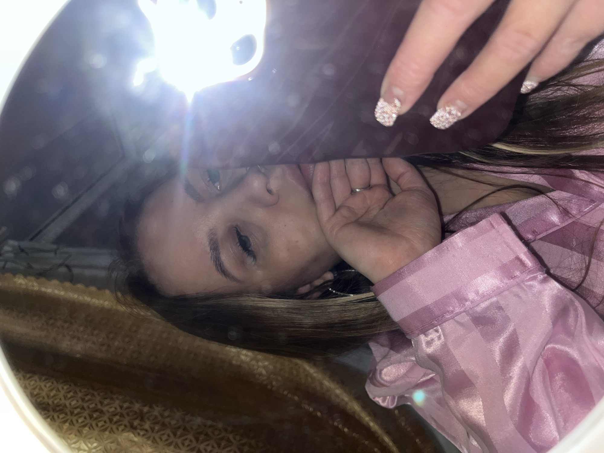 A woman with light brown hair and wearing a light pink, satin pajama top is looking into a circular mirror. The mirror is reflecting her face, hand, and the camera (or phone) she is holding. The woman's right hand is near her face, with a ring visible on her ring finger. Her nails are manicured with a glittery design. The mirror's frame appears to be white, and a gold, textured material is visible in the background of the reflection. There is no information to identify the location in Novopokrovskaya, Russia.