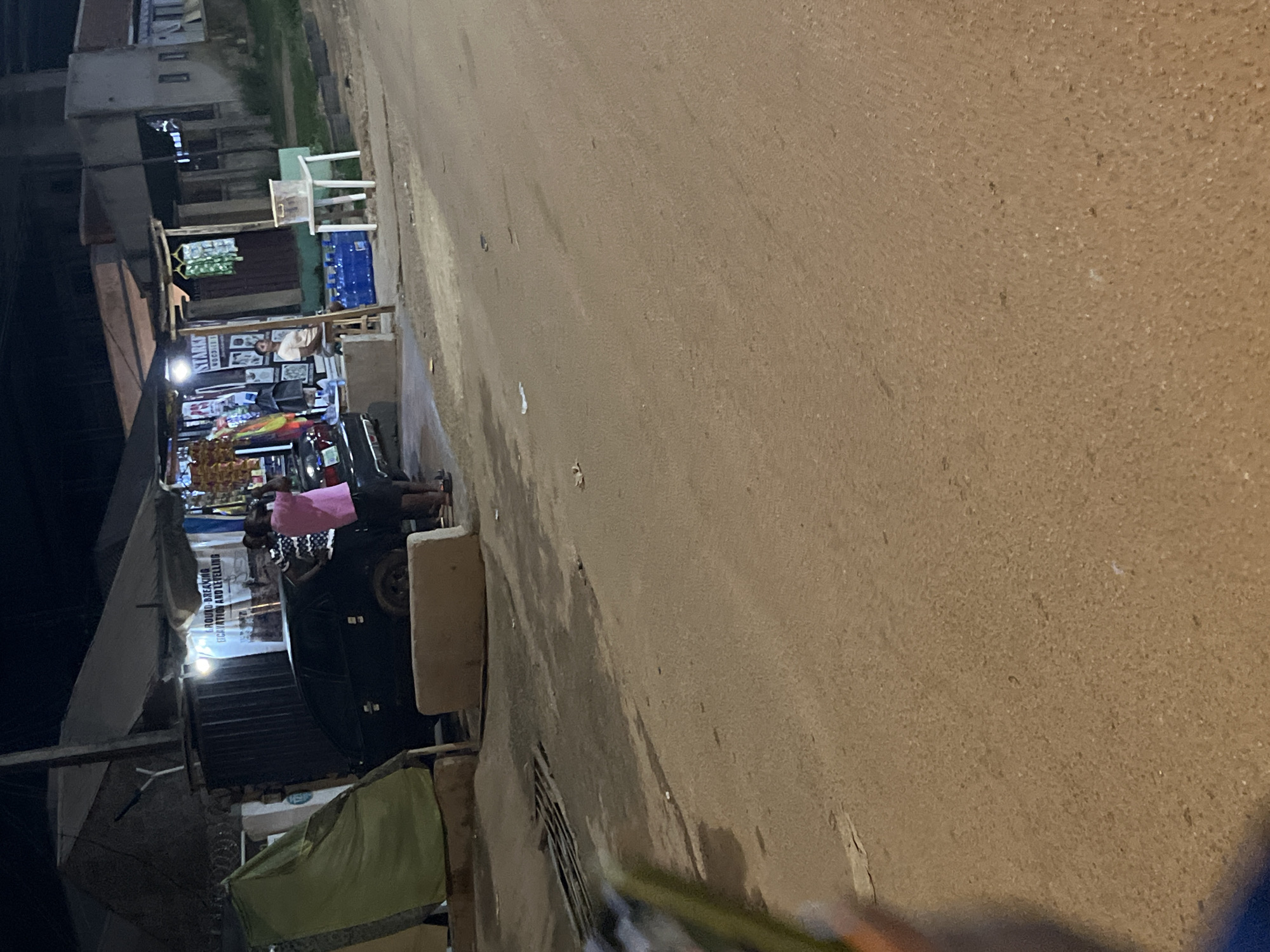 In Abuja, Nigeria, at nighttime, two individuals are standing near a black car in front of a roadside stall. One person, wearing a pink shirt and dark shorts, is holding a phone. The other person, in a striped shirt, is looking towards the first person. The stall has merchandise displayed, including packaged goods. Adjacent to the stall, there is a white table with a plastic container and blue bottled water. The street in front of the stall appears to be made of packed dirt. The scene is illuminated by artificial lighting, casting shadows and creating a dimly lit atmosphere.