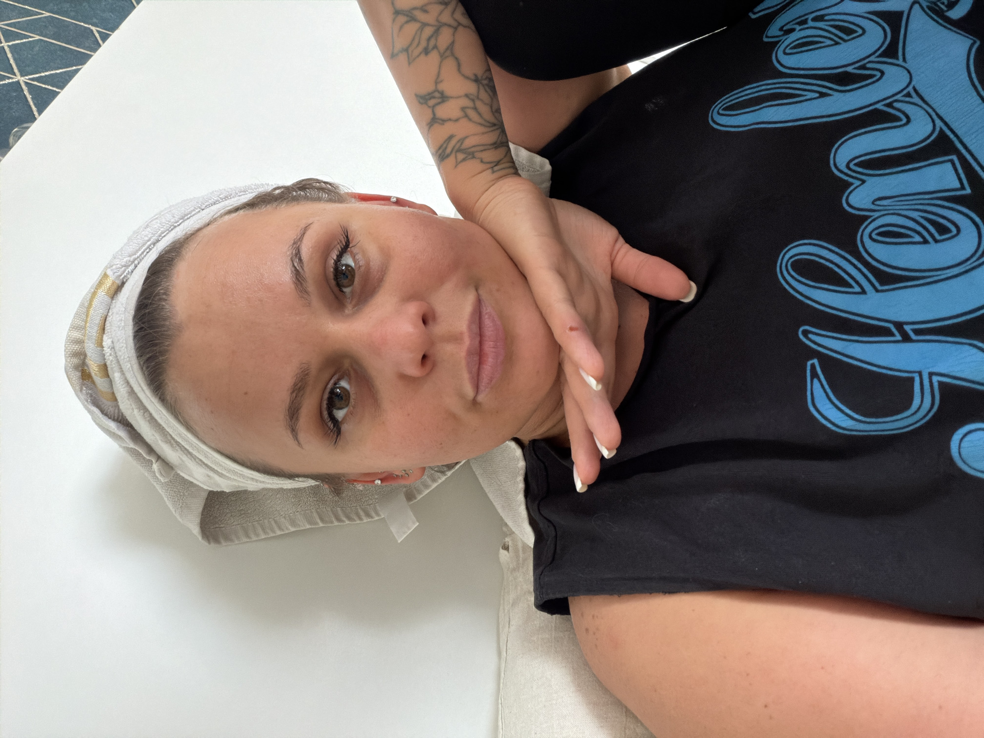 A female individual is depicted in a close-up, head-and-torso selfie, situated indoors within the city of Sarapul, Russia. Her head is wrapped in a light-colored towel with subtle striped accents, suggesting recently washed hair. Her facial expression is neutral, with direct eye contact to the camera. Brown eyes are visible with enhanced eyelashes. The individual wears a dark-colored sleeveless top featuring a light blue graphic, part of which reads "Henley" in a stylized font. Her right arm is bent, with her hand supporting her chin, displaying nails with white tips. A small red mark is visible on the back of her right hand. A linear floral tattoo is present on her right forearm. The left ear shows multiple piercings, including a stud and several small hoops. The background consists primarily of a plain white wall, with a small portion of a blue tiled surface with a geometric pattern visible in the upper right. A light-colored, textured fabric, likely a cushion or pillow, is visible on the left side and beneath her left arm.