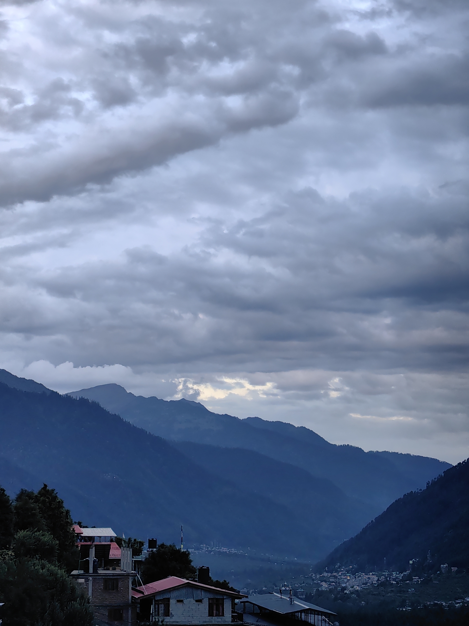 The image depicts a panoramic view of the village of Vashisht in Himachal Pradesh, India, as evening approaches. The sky is dominated by heavy, dark grey clouds, suggesting an impending storm or the remnants of one. Below the dramatic sky, a series of layered mountains recede into the distance, their slopes a deep, muted blue. The terrain is rugged and mountainous, typical of the Himalayas.
In the foreground and mid-ground, several houses are visible, built with a mix of brick and concrete. Some have red tile roofs, and others appear to be under construction or unfinished. The architecture suggests a rural or semi-urban setting. The houses are nestled on the slopes, interspersed with dark green trees, likely pine or deodar, which are common in the region.
Further into the valley, a small town or village is illuminated by artificial lights, creating a cluster of warm glows against the darkening landscape. This settlement is situated between the mountains, hinting at the presence of a river or valley floor. There are no visible people or animals in the frame. The overall atmosphere is serene yet imposing, characterized by the grandeur of the mountains and the brooding sky. The lighting conditions indicate a low-light environment, possibly due to cloud cover and the approaching twilight.