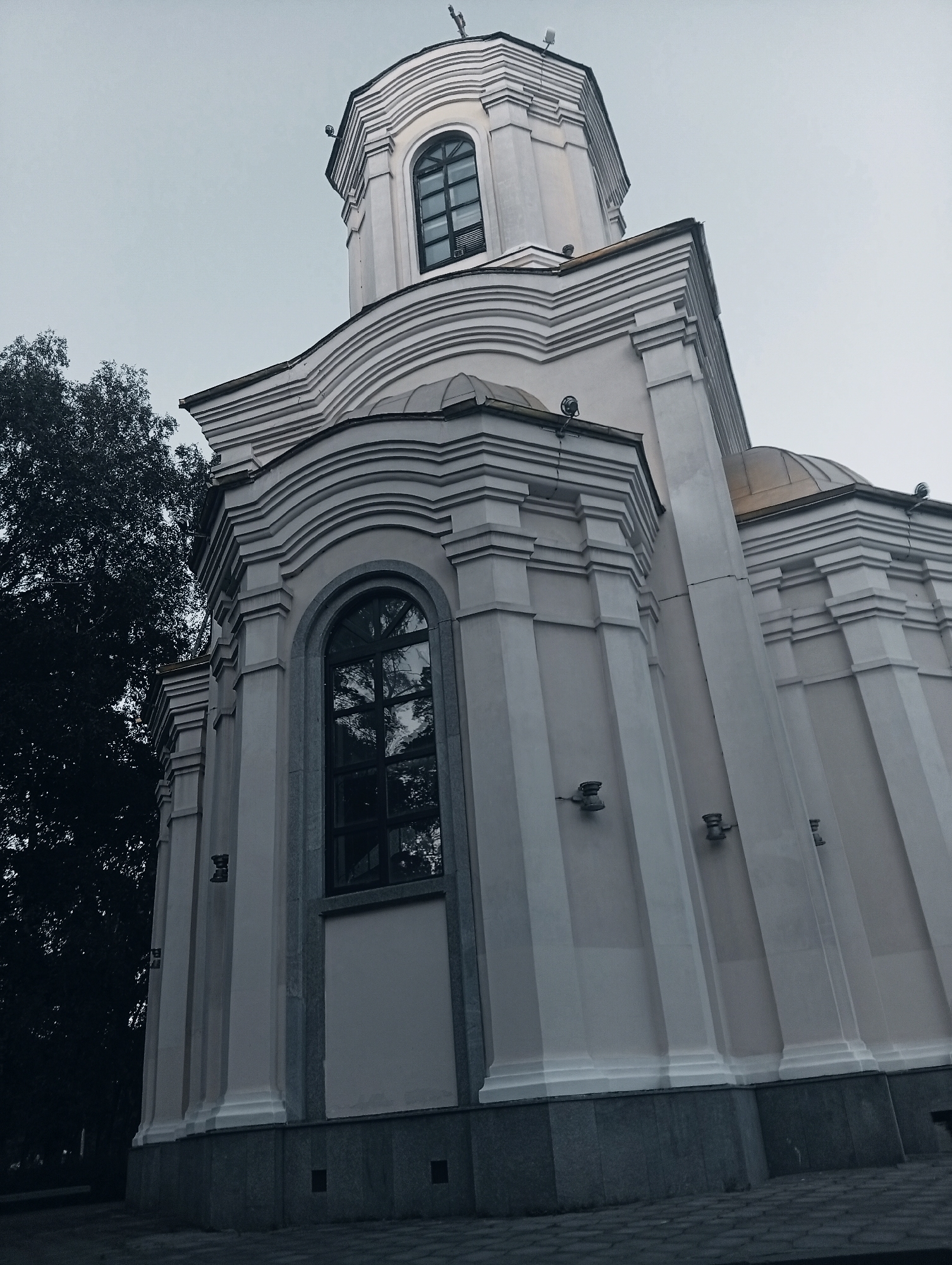 A low-angle, medium shot captures the side and dome of a light-colored, European-style church against a pale, overcast sky. The church features architectural details such as arched windows with dark panes, decorative moldings along the roofline, and vertical fluting on its exterior walls. A large, dark tree with dense foliage is visible on the left side of the frame, partially obscuring the church's left flank. The ground in the foreground is paved with cobblestones. The image is dominated by muted tones of gray, white, and dark green, with a hint of gold on the dome.