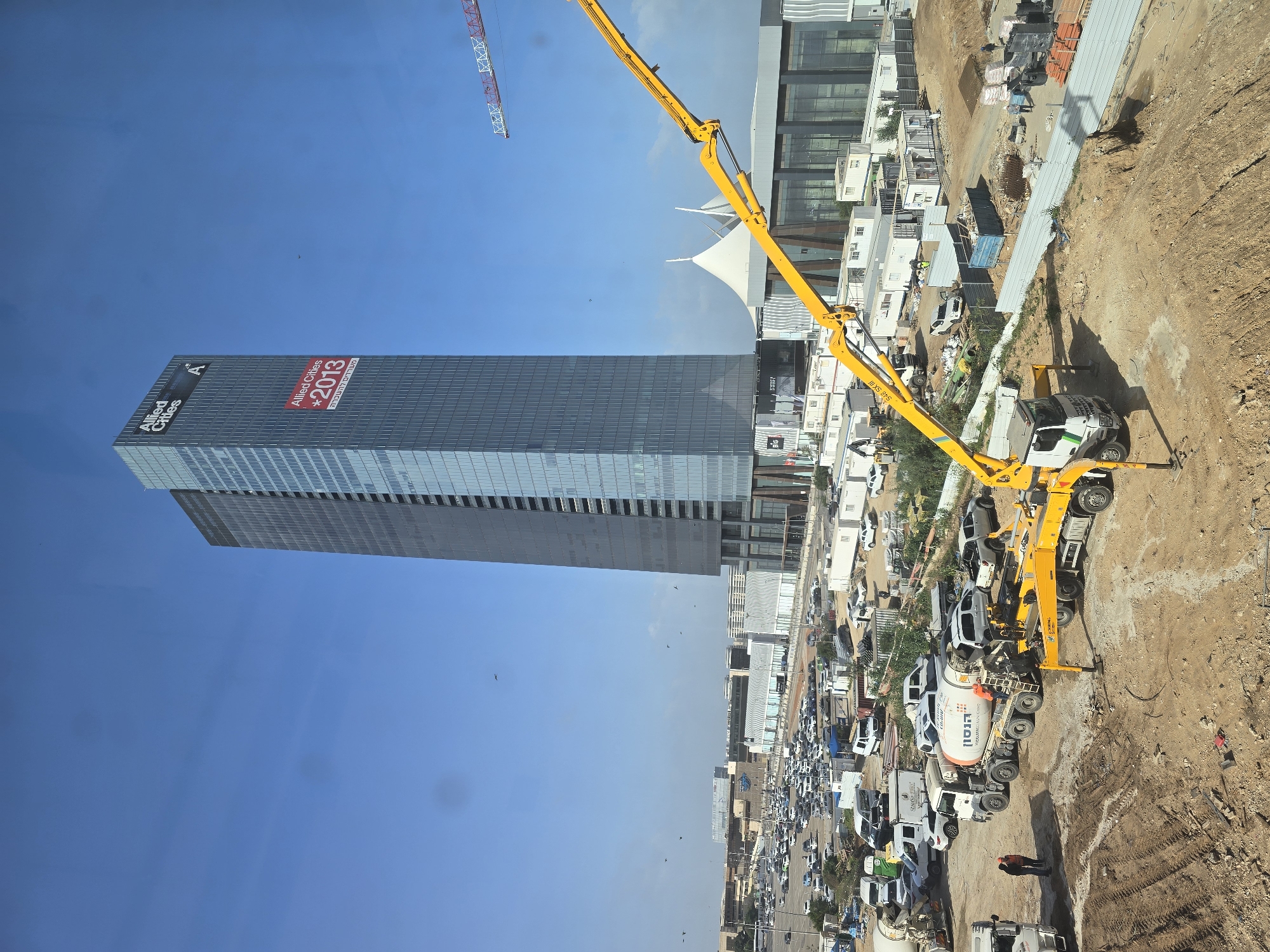 The image shows a construction site with a large tower in the background. The tower is labeled "Allied Cities" and has a sign indicating "2013". A yellow construction crane is positioned near the building, and several construction vehicles, including concrete mixers, are visible on the ground. The environment appears to be a busy urban area, with other buildings and vehicles in the distance. The sky is clear and blue. The image is taken from a high vantage point, possibly from another building or elevated structure.