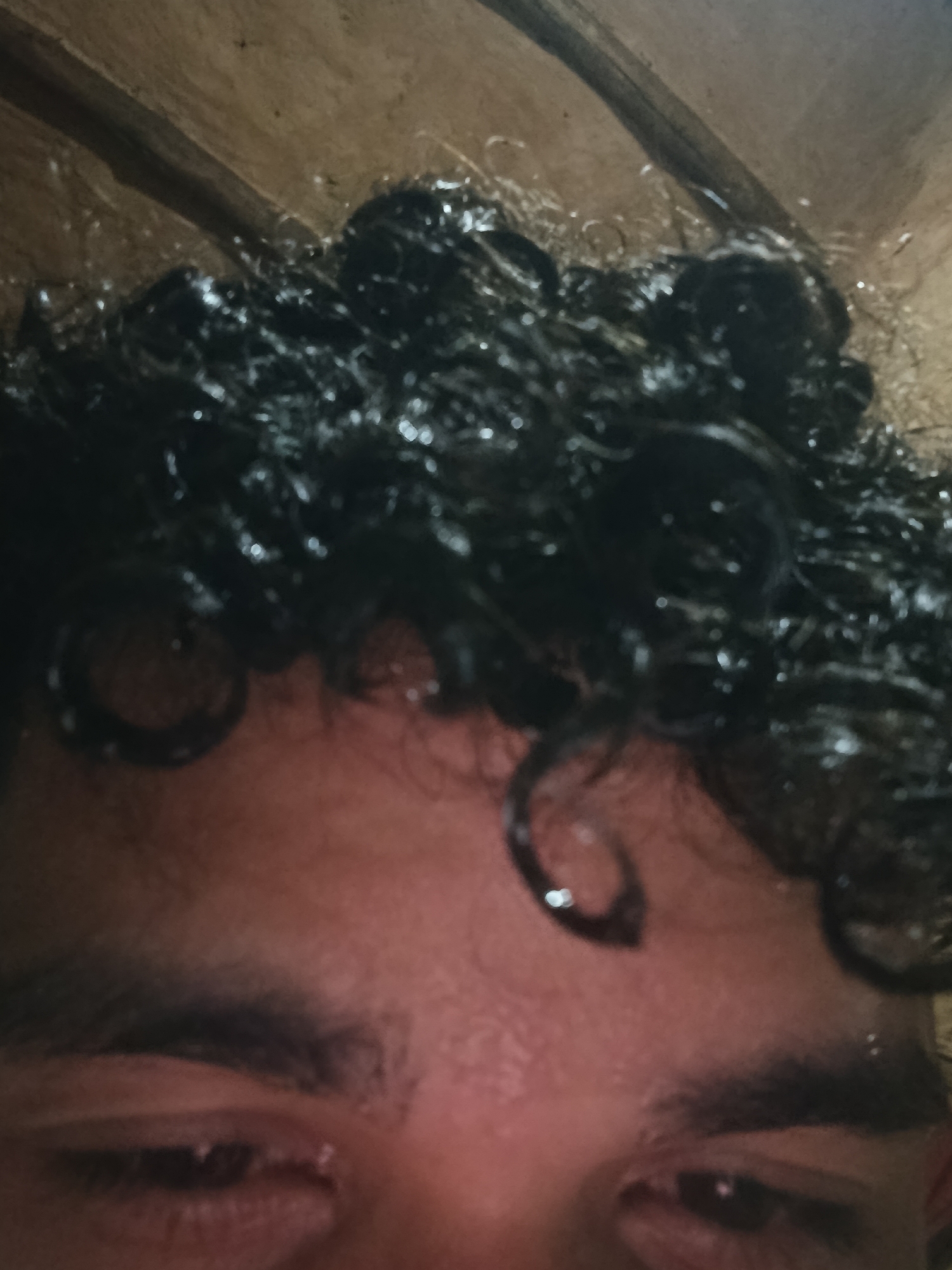 A close-up shot shows the face of a person with wet, dark curly hair covering their forehead. The person's eyes are visible, looking slightly downward. The background appears to be a tan or brown, possibly a ceiling or roof. There is no information in the image to identify the location as Juan Griego, Venezuela. The environment appears to be indoors.