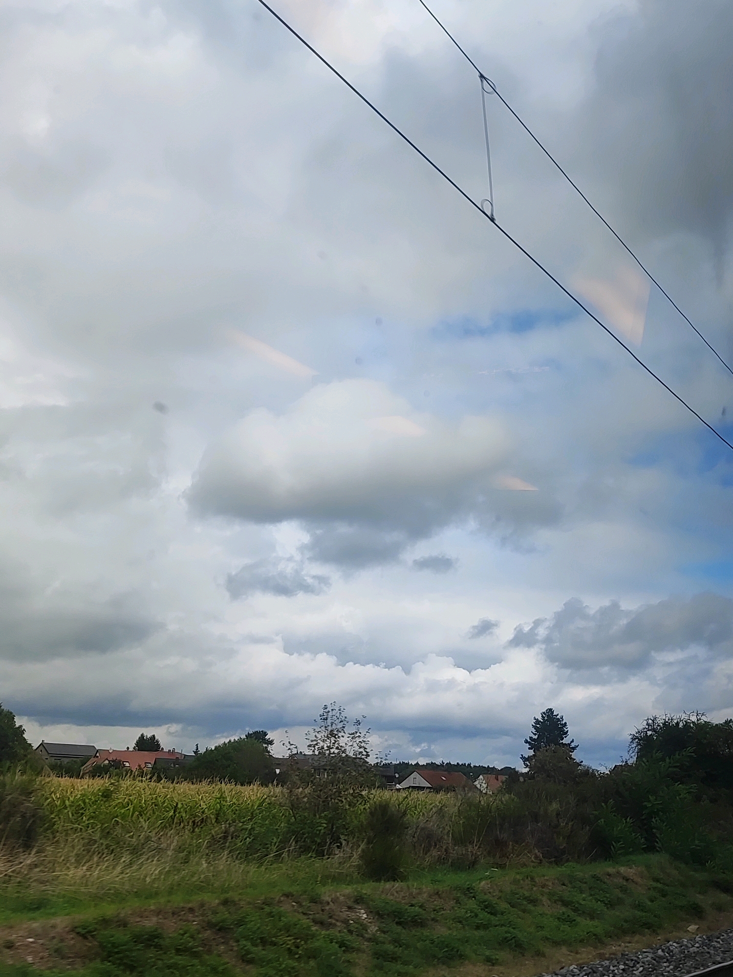 The image depicts a rural landscape under a cloudy sky, as viewed from a train. In the foreground, there is a grassy embankment with a gravel track to the right, likely railroad ballast. Beyond the embankment, a field of tall green crops, possibly corn, stretches across the midground. Several buildings, including houses with red-tiled roofs and other structures, are visible behind the crop field, nestled amongst trees. A dense line of trees forms a backdrop to the settlement. The sky is filled with a mix of dark and light clouds, with patches of blue visible. Two dark overhead wires, characteristic of an electric railway, bisect the upper portion of the image, with a clear plastic insulator visible between them. There are no people or animals visible in the scene. The overall impression is one of a pastoral setting in the German countryside.